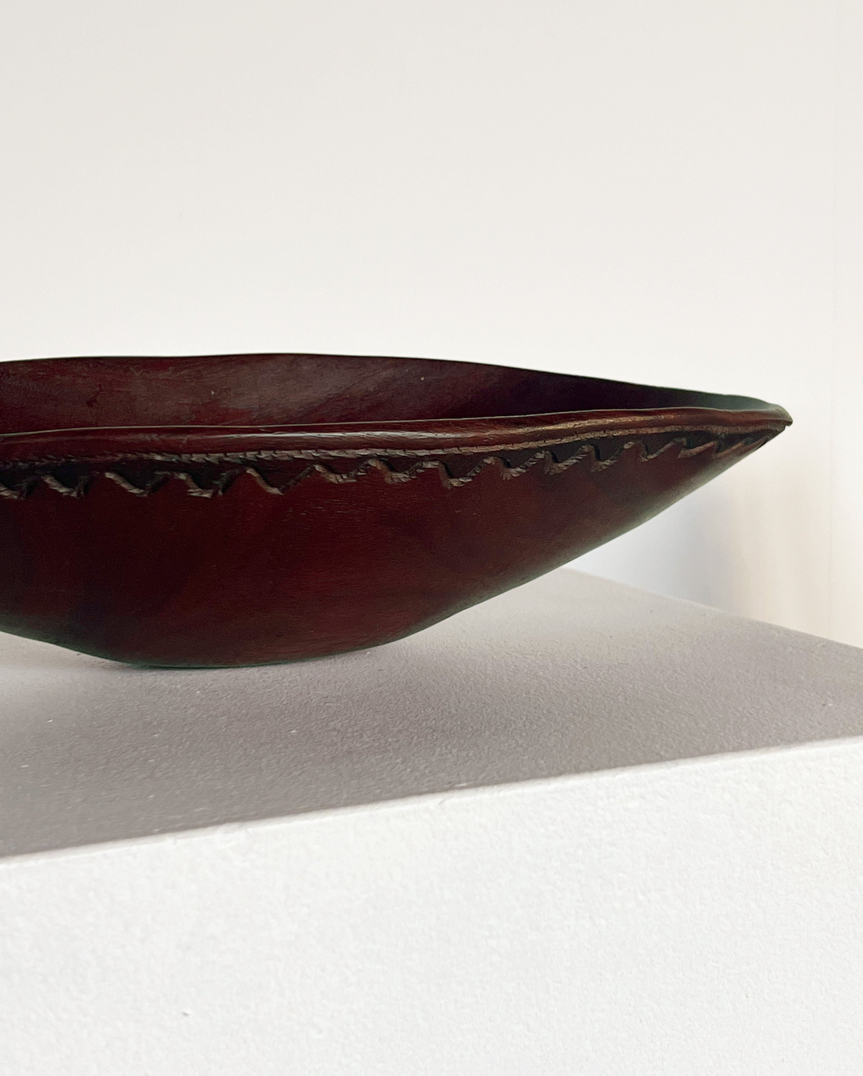 The Carved Wooden Bowl by Porter Studio, made from handcrafted mahogany with a dark finish, rests on a white surface. This artisan product showcases an intricate zigzag carved pattern along the rim, adding detailed artistry to its simple design. The hand-carved texture elevates its natural wood appeal.