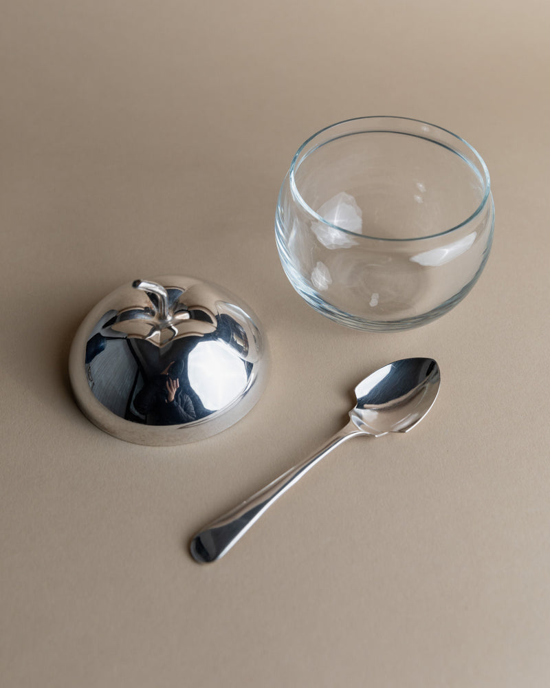 On a smooth, neutral surface lies the "Silver & Glass Apple Jar" by Bottega Jacobs, featuring an apple-shaped, shiny silver-plated lid with a leaf-shaped handle and a matching spoon. The reflective lid adds an artistic touch by mirroring its surroundings.