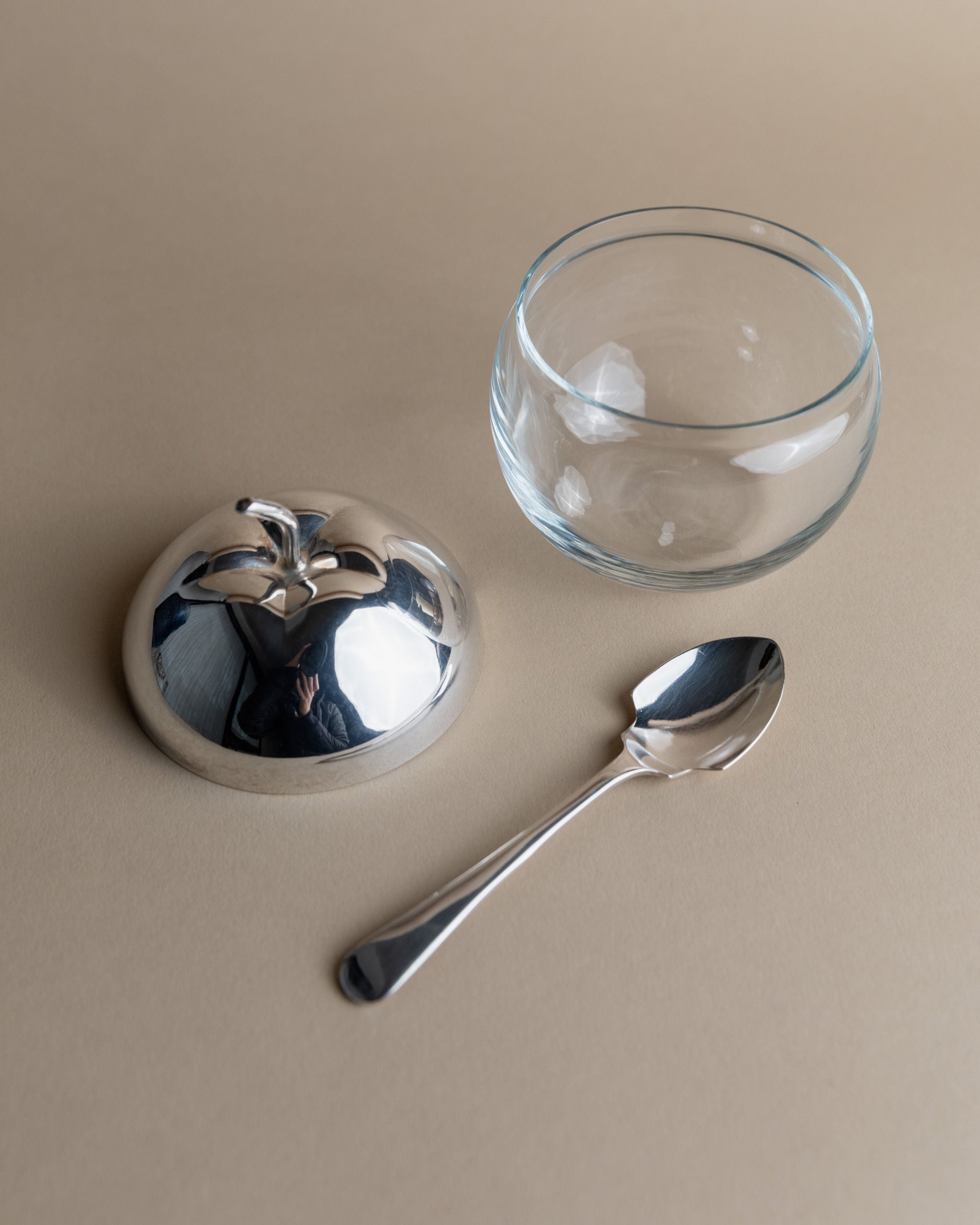 On a smooth, neutral surface lies the "Silver & Glass Apple Jar" by Bottega Jacobs, featuring an apple-shaped, shiny silver-plated lid with a leaf-shaped handle and a matching spoon. The reflective lid adds an artistic touch by mirroring its surroundings.