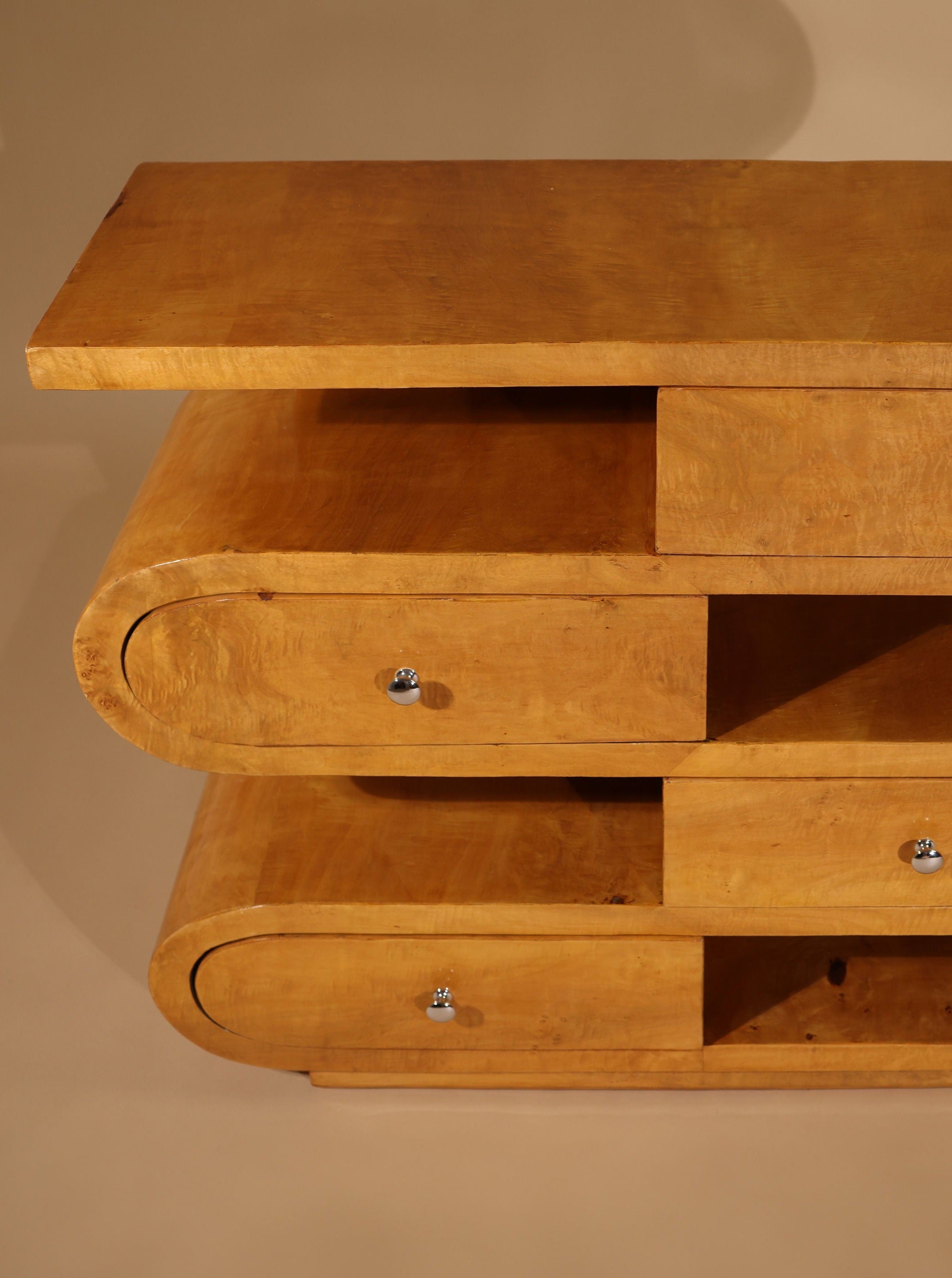 A mid-century design Hebi Drawers Console Burl Wood by Meola Interiors, this wooden piece of furniture features three tiers. The top tier is a flat surface, while the two lower tiers have curved ends and small drawers with silver knobs. The light, natural finish is accentuated by the elegant burl wood veneer.