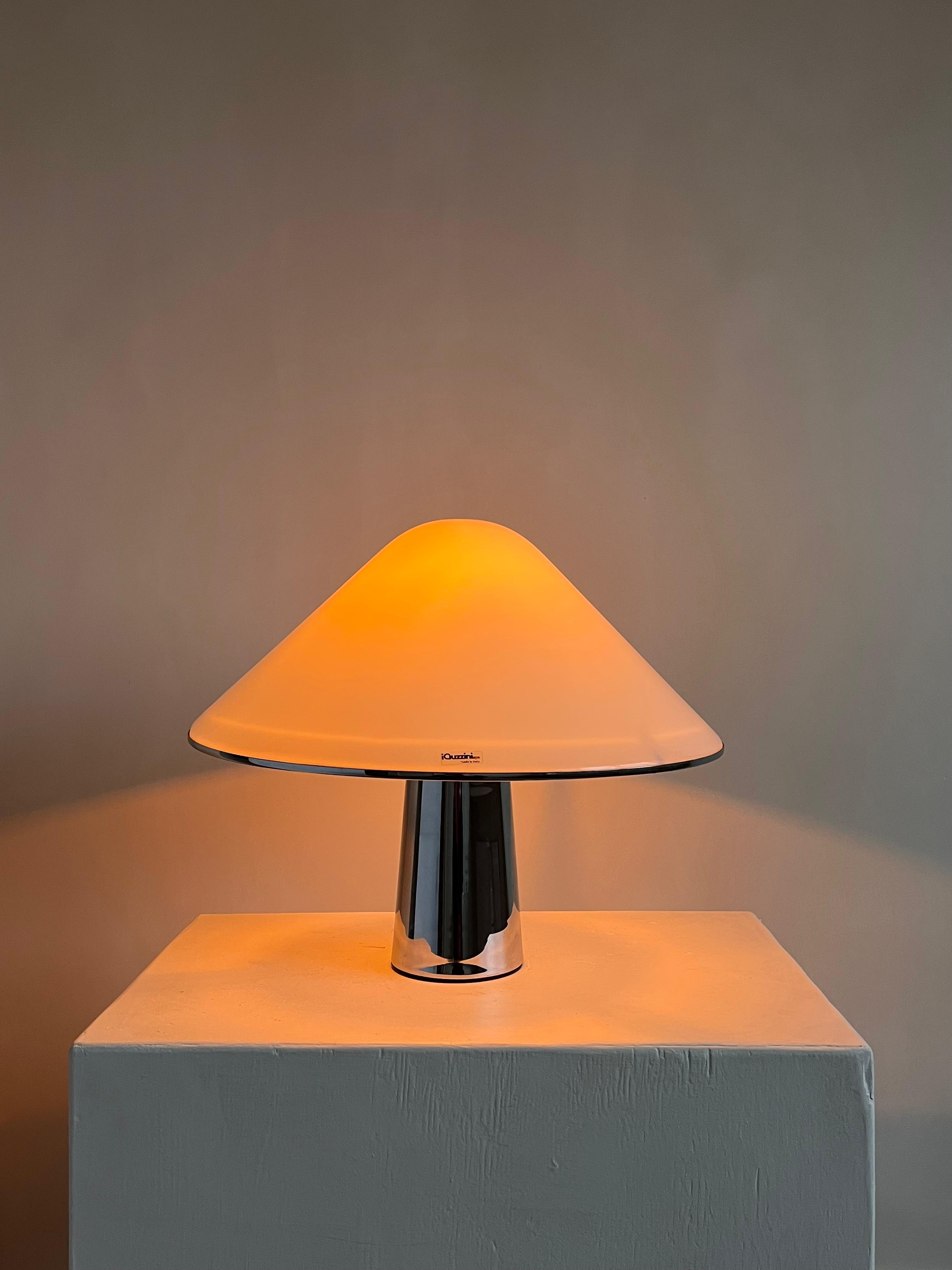 The Harvey Guzzini Elpis Table Lamp by septembre studios, inspired by Luigi Massoni, features a sleek dome shade and metallic cylindrical base that emits a warm glow atop a minimalistic white pedestal, complementing its vintage condition against a plain gray background for cozy ambiance.