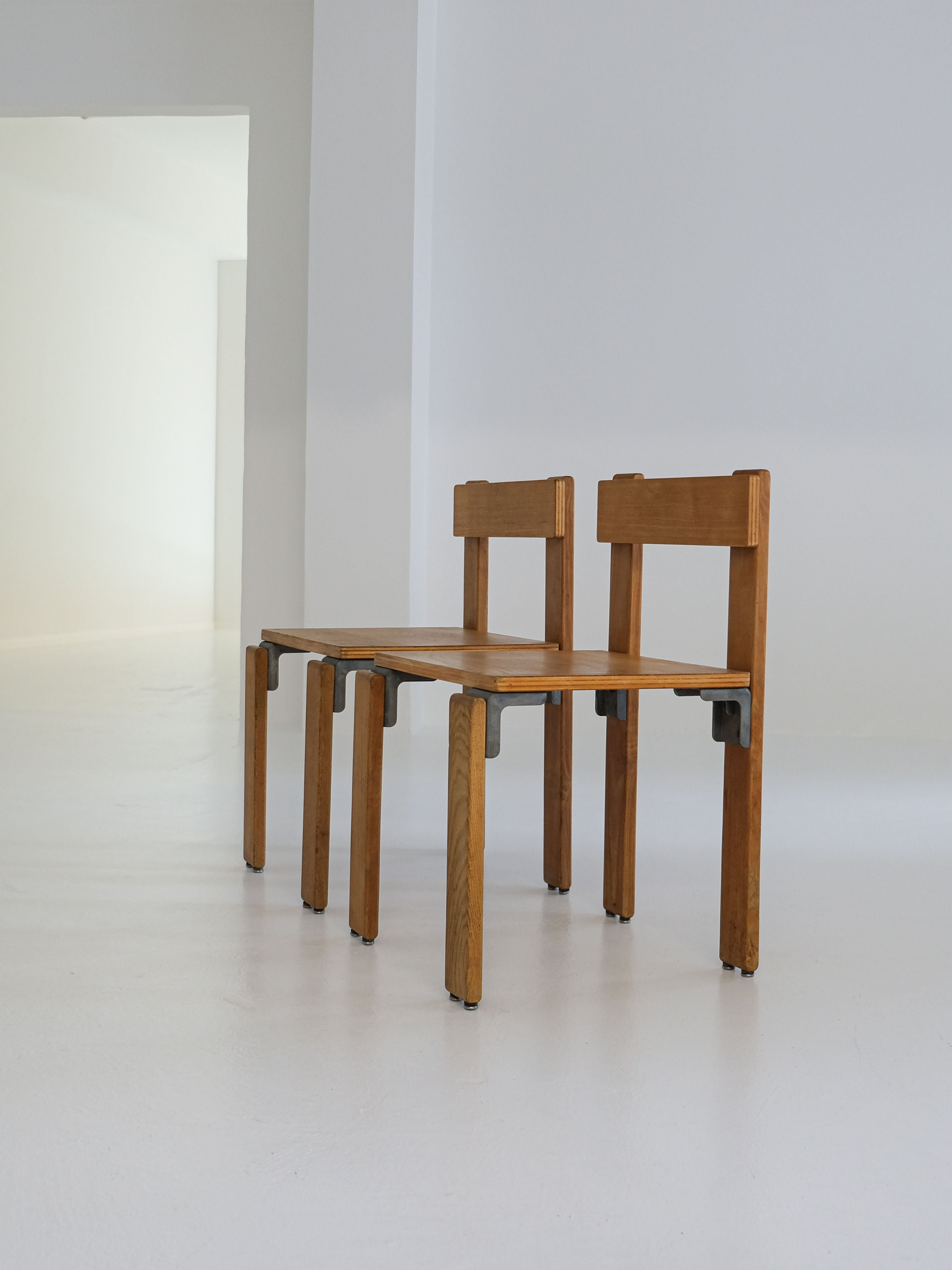 The "Les Carats" dining chairs by George Candilis and Anja Blomstedt, 1970s, from antibeige, are minimalist wooden chairs crafted from oak plywood with straight backs and seats interconnected in a sleek design. They rest on a smooth, light-colored floor against a plain white wall.