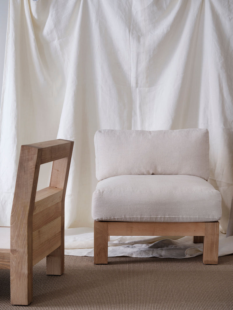 The "Forma" Armchair by FEBRERO SHOP, made of solid wood with a cushioned seat, is set on a beige rug alongside its matching chair. Draped white fabric in the background adds an elegant touch.