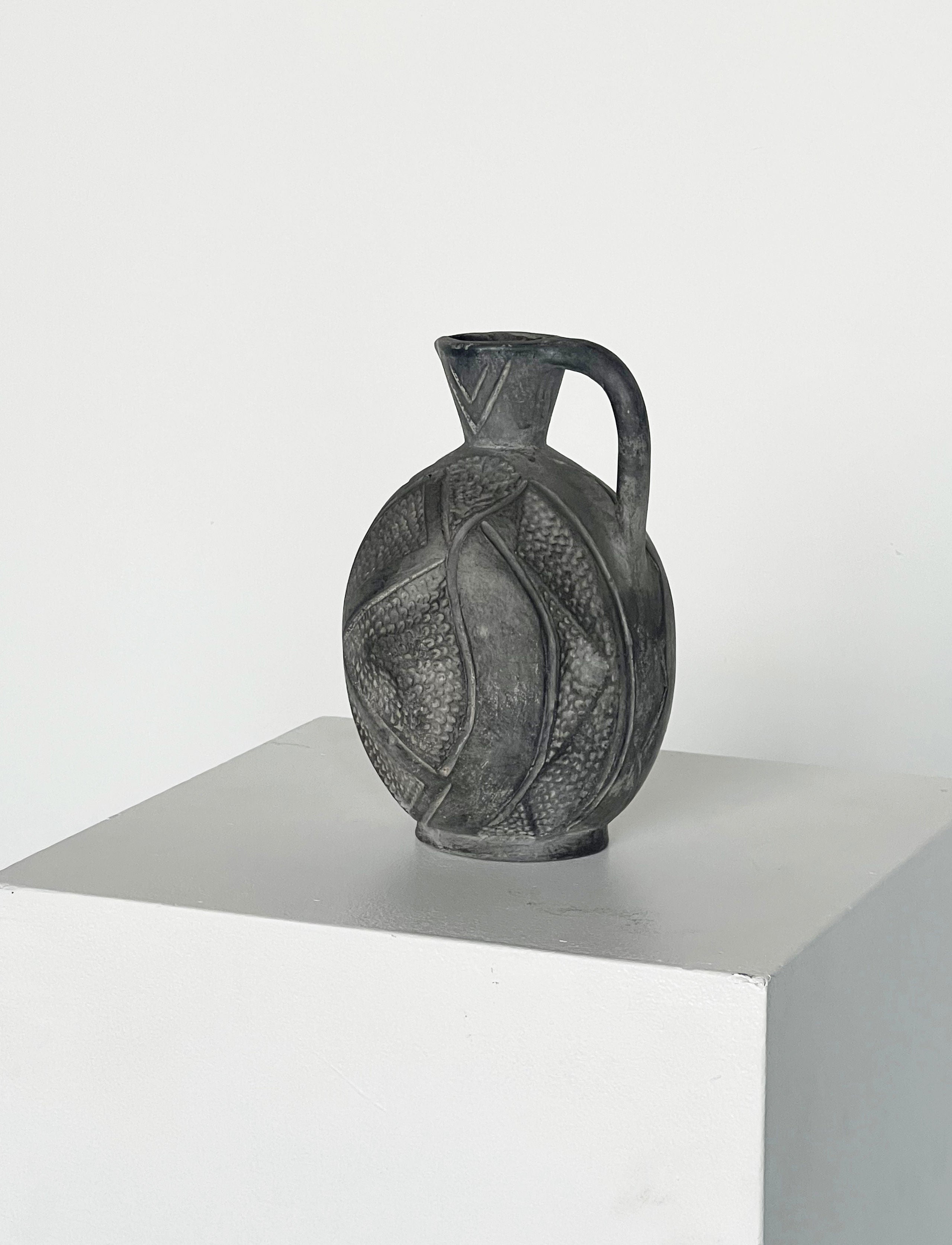 A round, dark handmade jug from Porter Studio, featuring hand markings and a textured swirling pattern, elegantly sits atop a white pedestal against soft grey tones. The Jug with Hand Markings boasts a slender neck and handle, showcasing intricate craftsmanship.