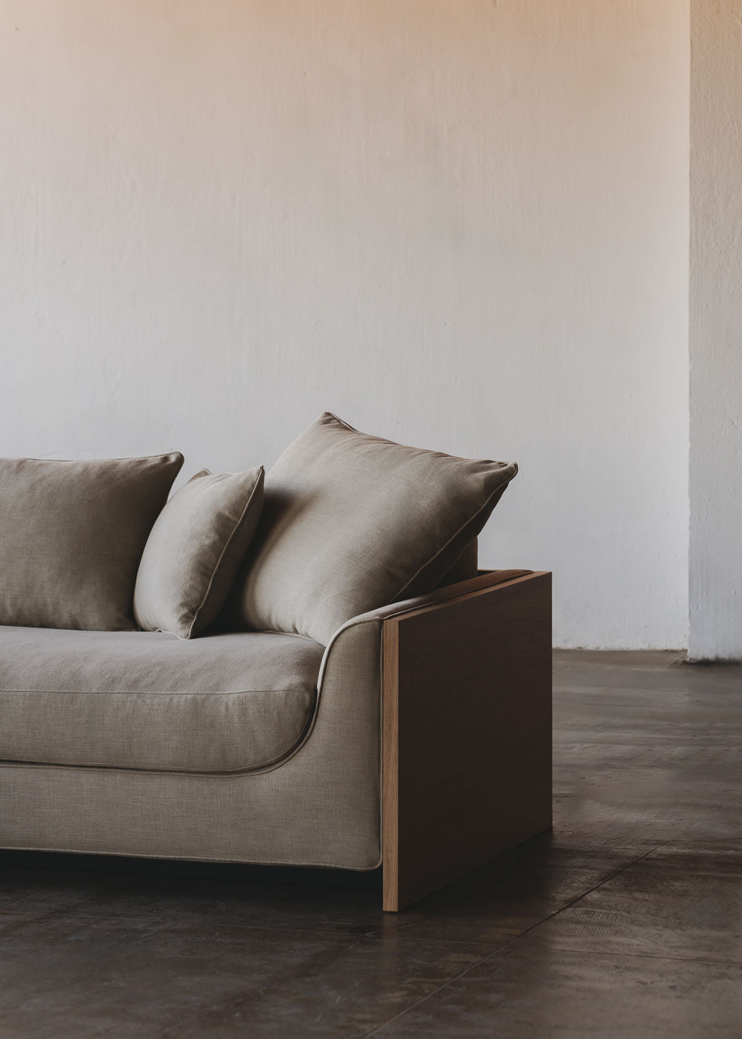 A Marina Sofa by ASERIES OF OBSTACLES, SL with light brown cushions is placed against a minimalist off-white wall. It features a solid oak frame and removable backrests and sits on a polished concrete floor, enhancing the stylish and contemporary living space.