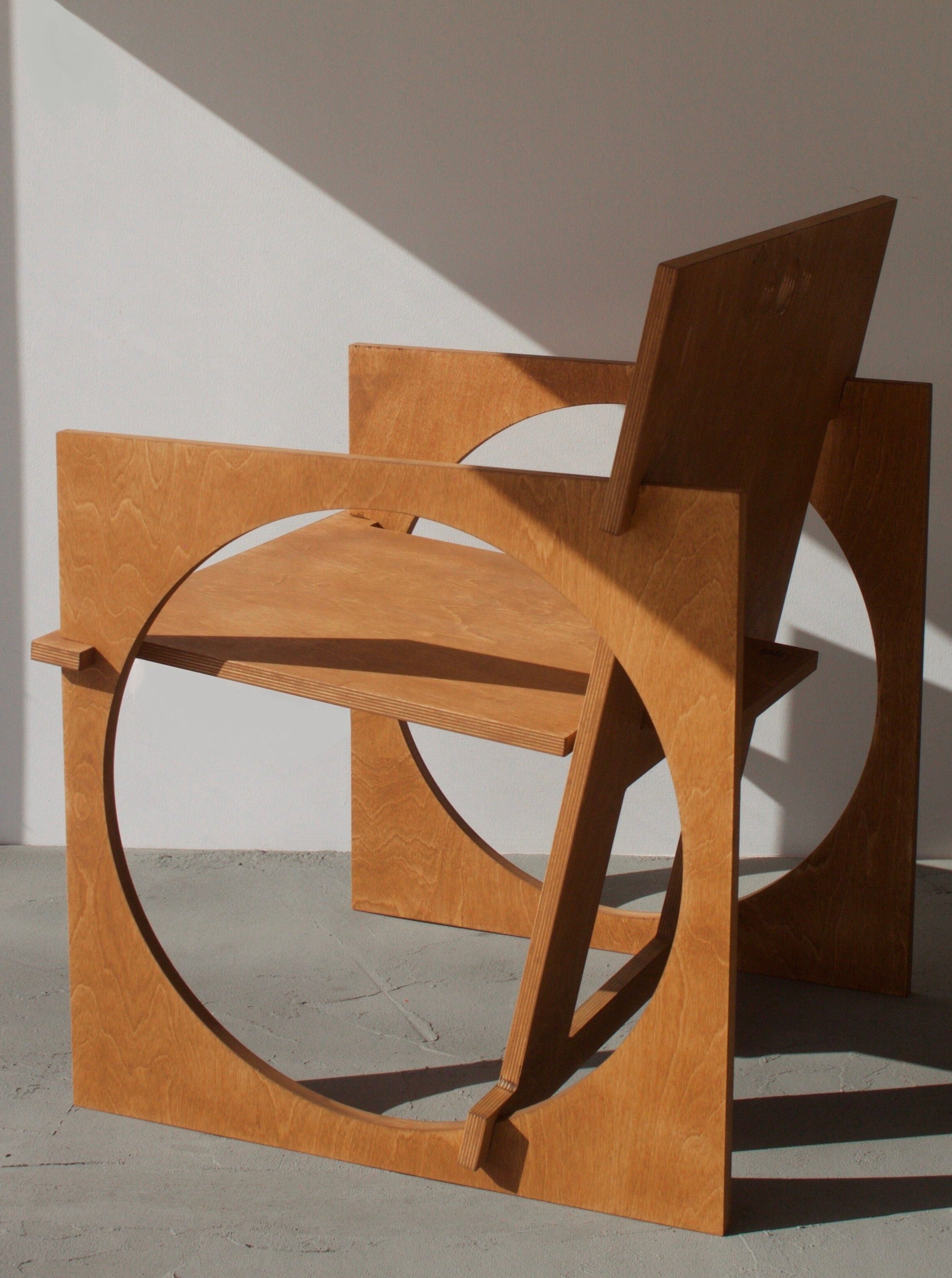 A modern wooden chair, known as the Edoardo Lietti Studio Sedia Tonda, with a unique circular design on its side, set on a concrete floor against a white wall, illuminated by natural light casting shadows.