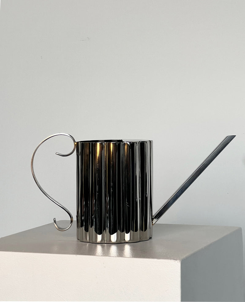 The Fluted Watering Can by Porter Studio, featuring a ribbed cylindrical body, elegantly curved handle, and long, narrow spout, creates an ornate silhouette on a white pedestal against the plain grey background.