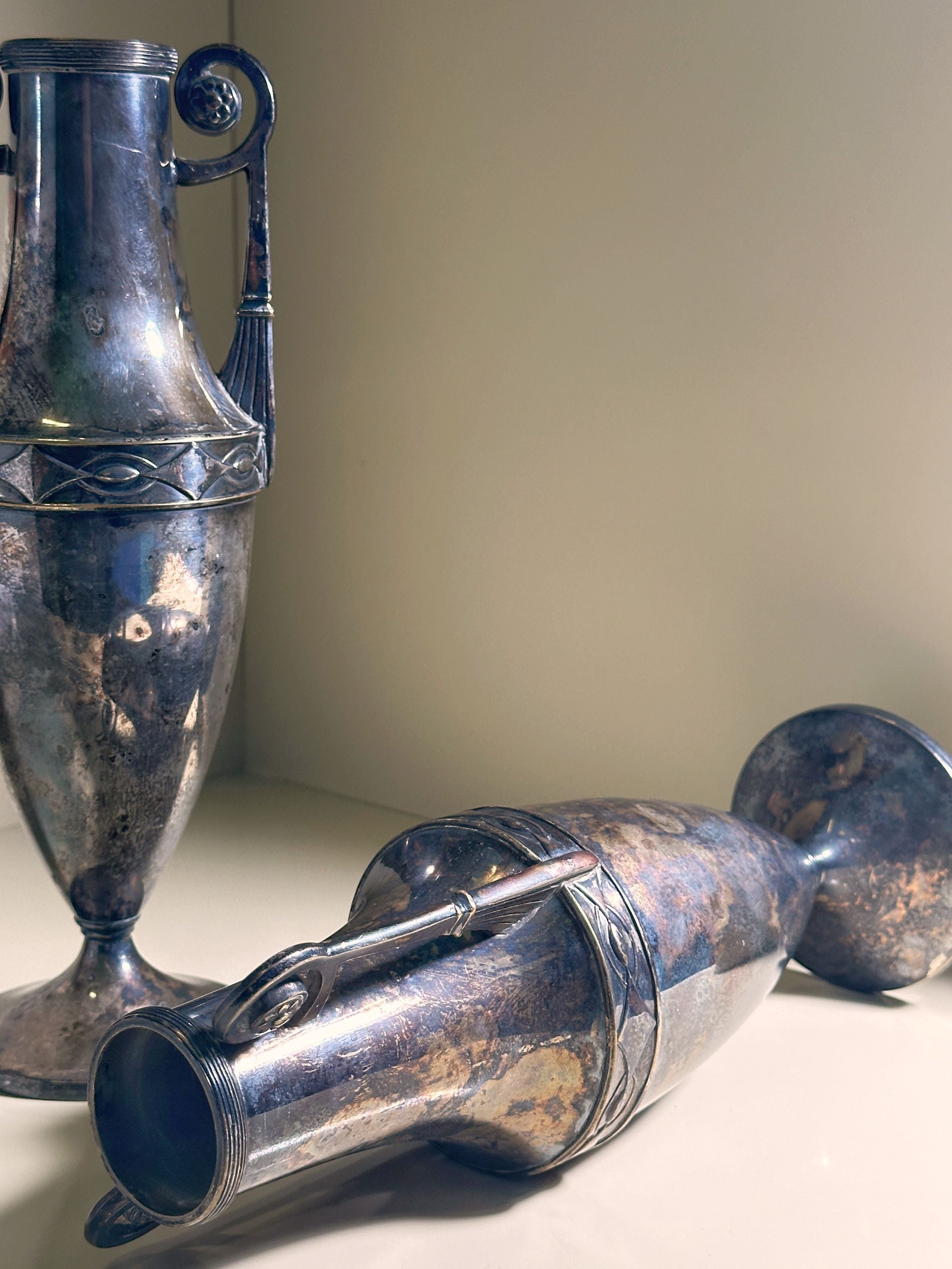 A pair of weathered silver-plated vases, reminiscent of Amphora design from 1890 by Dodo Vintage, features intricate band patterns and ornate handles—one vase upright, the other lying down against a neutral background.