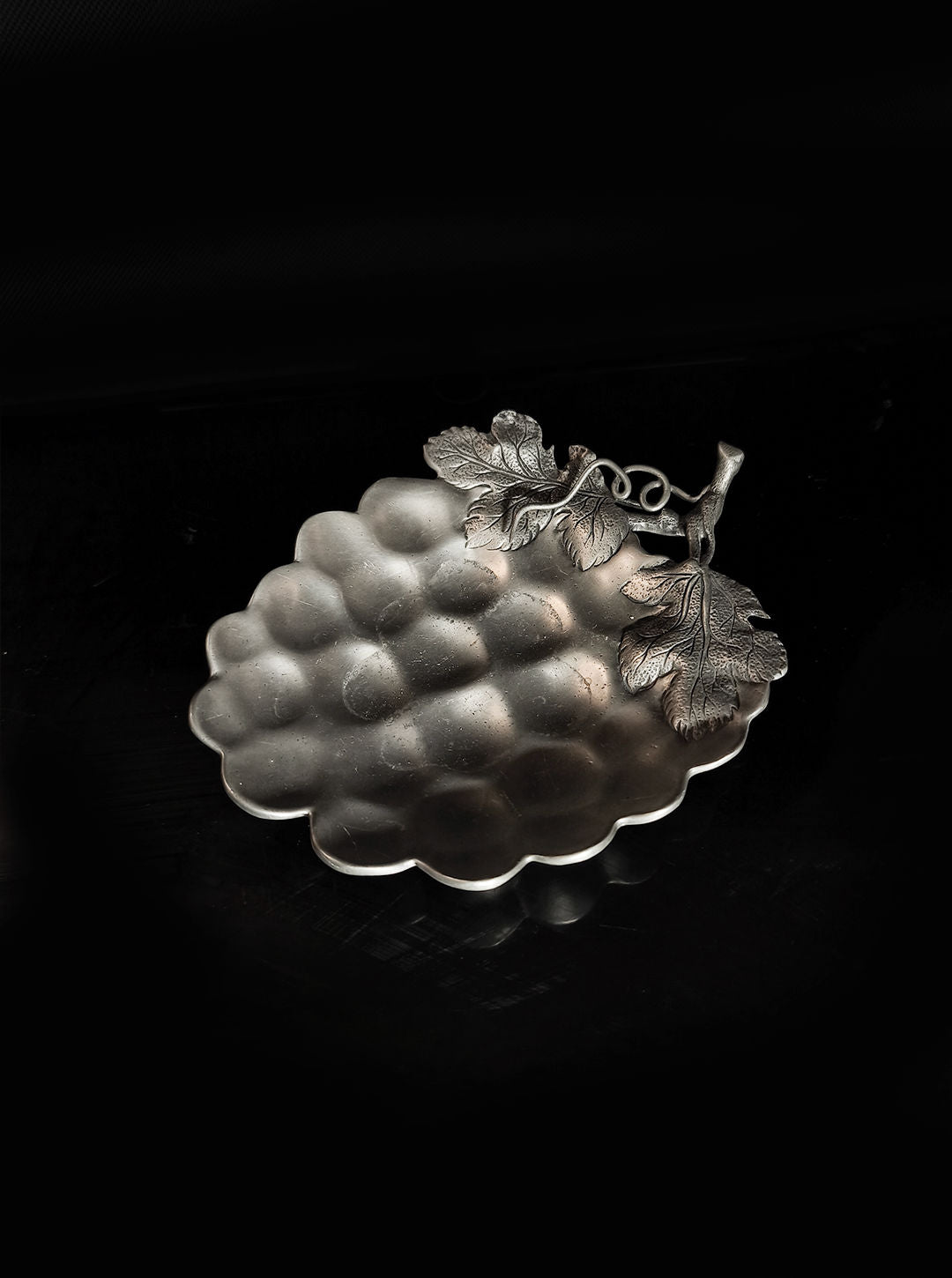 This Grape Vide Poches by Les Objoies is a pewter dish shaped like a grape, featuring an ornate handle with intricate leaf designs set against a dark silver background.