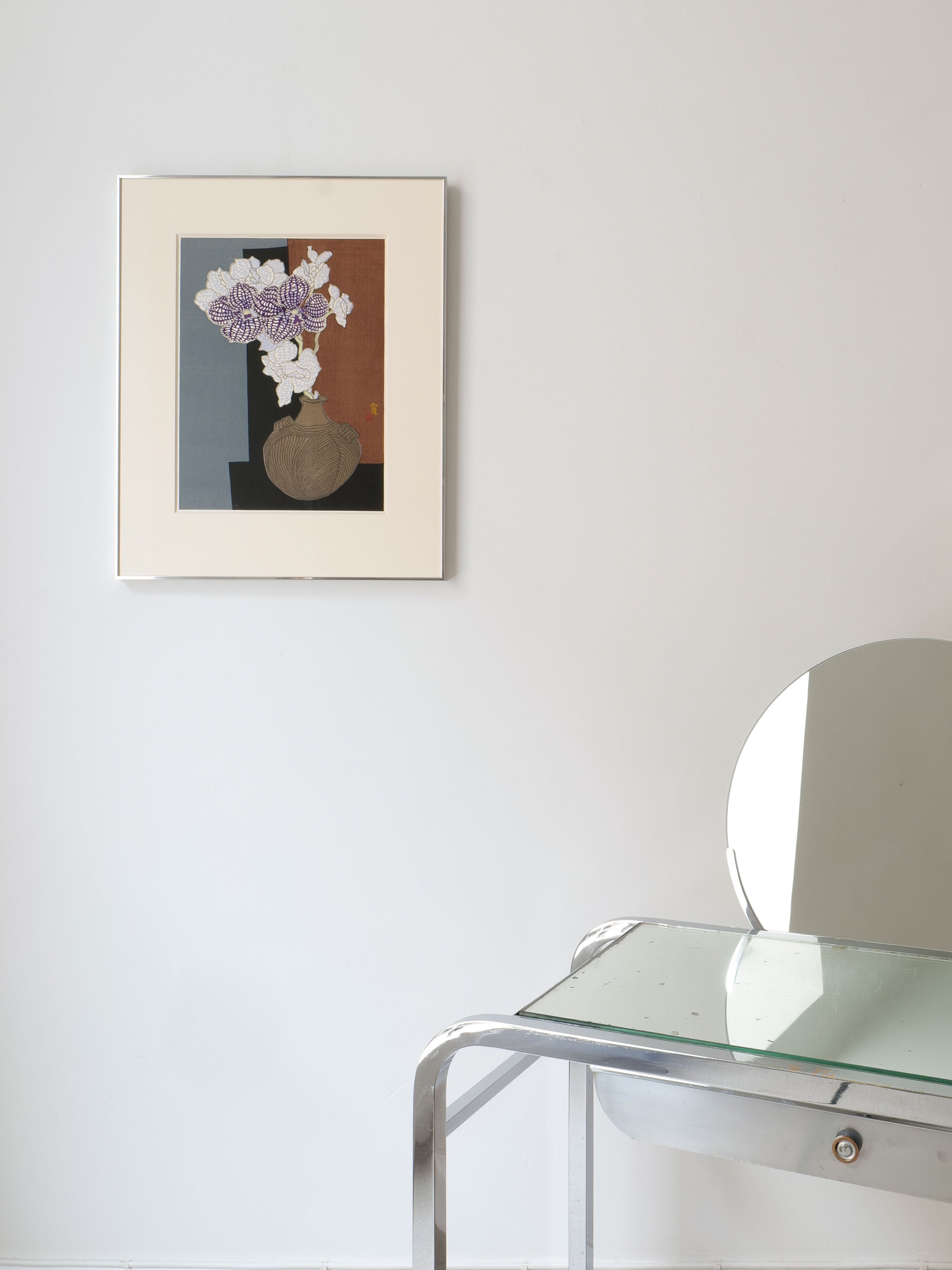 A minimalist room showcases the Orchid Woodcut Print Tokuriki Tomikichiro Japan 1980s from Collection apart on a white wall. Below, a sleek glass table pairs with a modern metal chair with a circular backrest, creating a clean and contemporary look.