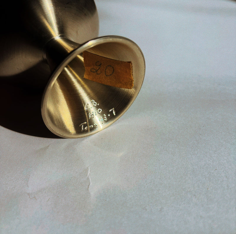 A Huerto brushed metal dessert coupe from a set of 4 rests on its side on a white surface. Its wide base features a yellow sticker with "20" handwritten and partially visible engraved text and numbers.