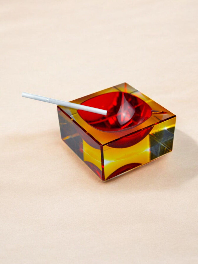 The Sommerso Ashtray by Seguso, a 70s Murano creation from Malamar Studio, sits on a light beige surface. This vibrant square collectible features red, yellow, and brown geometric designs with a sommerso glass technique; an elegant cigarette rests on its edge.