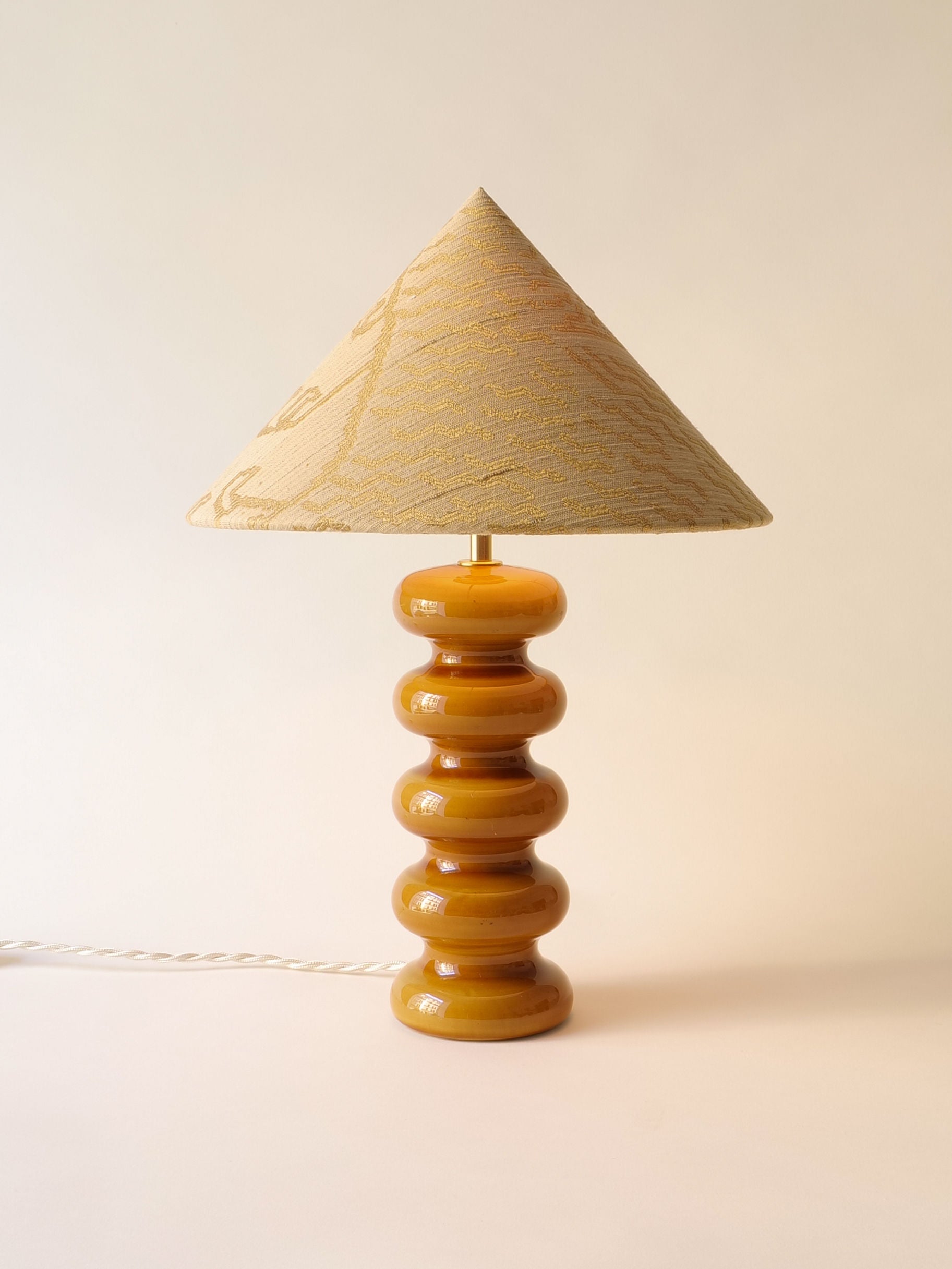 The Sculptural Ceramic Lamp by Collection apart features a geometric ceramic base with stacked, rounded shapes in shiny amber; it's adorned with a textured conical lampshade in subtle patterns on an off-white background, evoking Doulton England elegance. A white cord extends from the base.