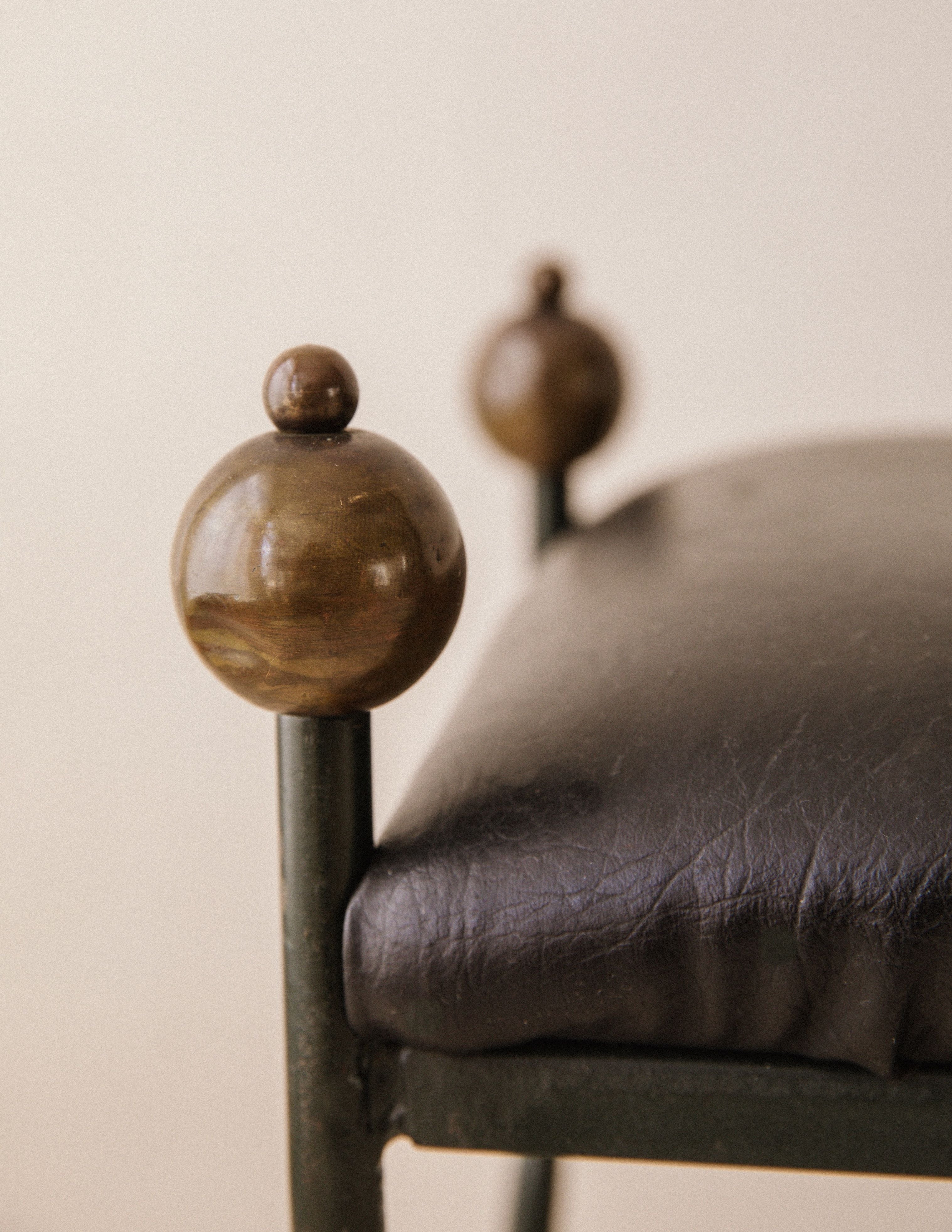 Close-up of the Arcā Collections Mid-Century Wrought Iron Bench, featuring a black leather seat and round brass accents on its metal frame, set against a neutral background.