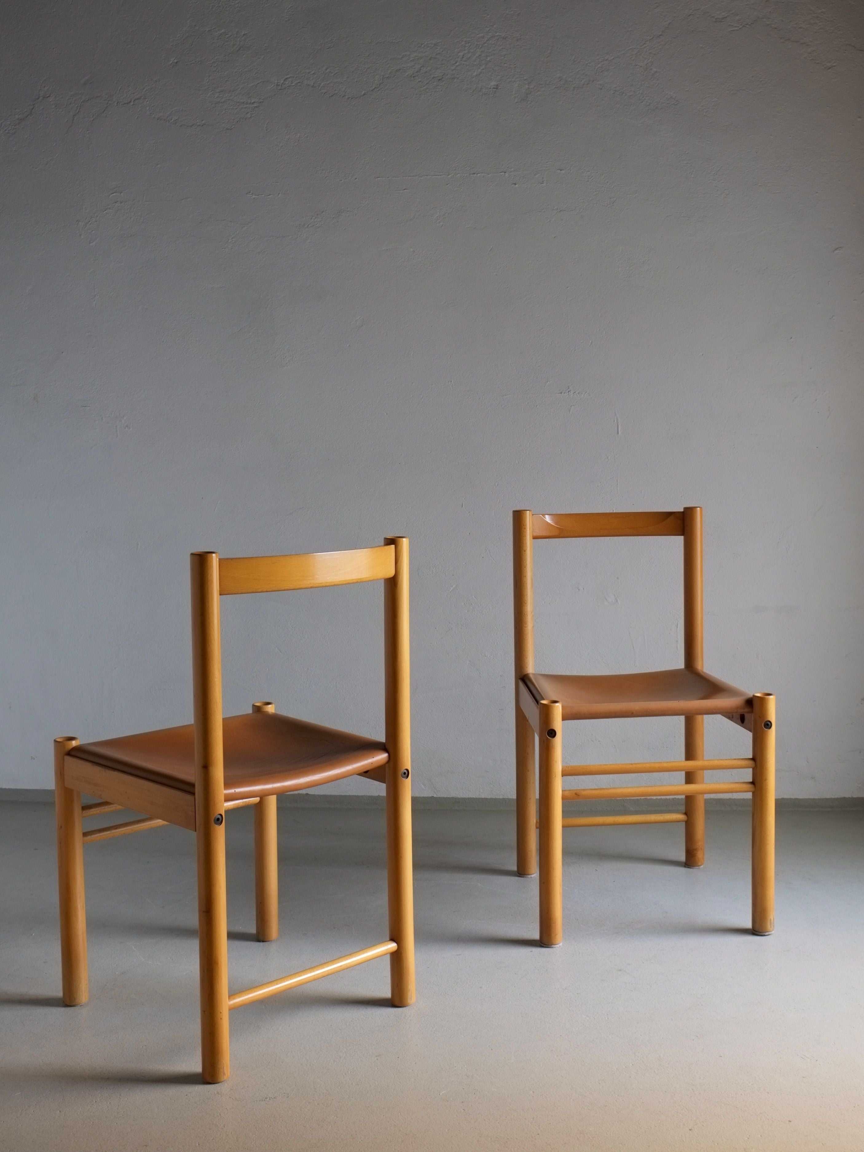 Two vintage-style dining chairs from Veter Vintage's "6 Beech Wood Cognac Leather Chairs from Ibisco, Italy, 1960s" collection are arranged on a plain surface against a gray wall. Crafted from beech wood, the chairs showcase a simple design with brown leather seats and are positioned at slight angles to each other, exuding a minimalist and timeless ambiance.