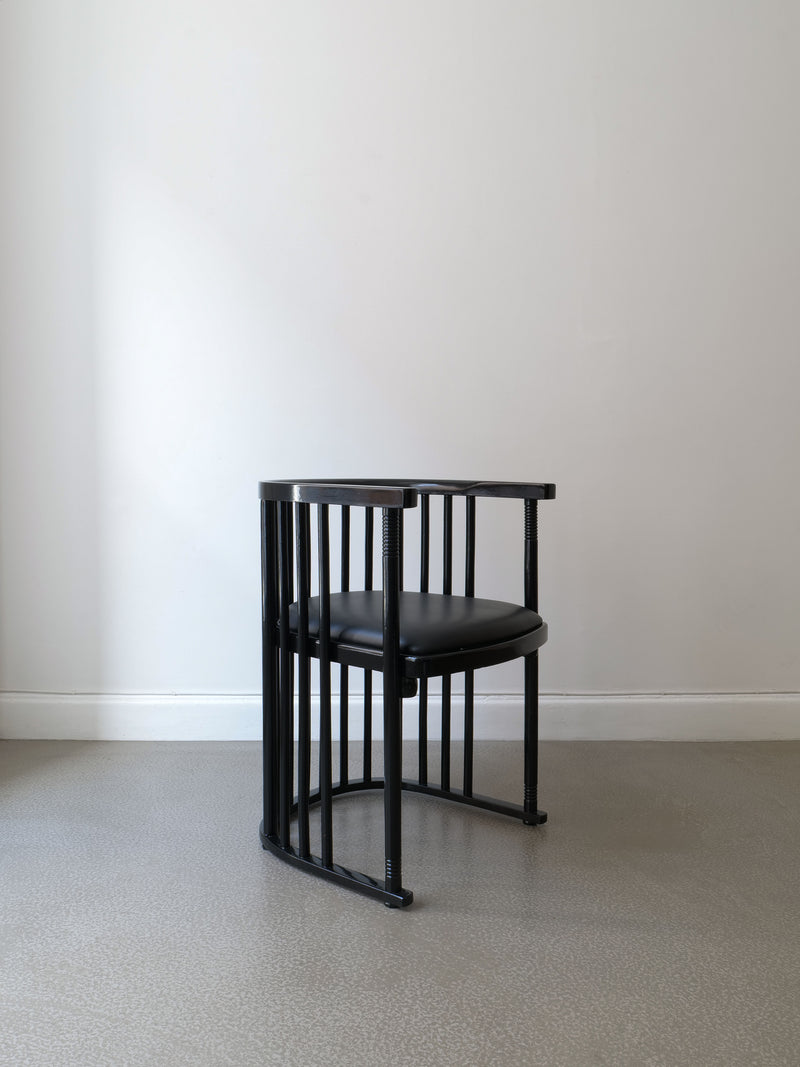 The Bentwood Armchair Josef Hoffman by Collection apart, featuring a modern black design with lacquered bentwood and a cushioned seat, is placed on a gray floor against a plain white wall.
