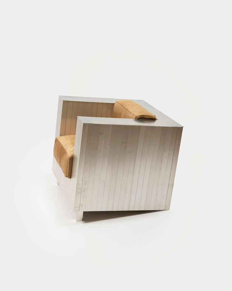 The Aluminum Armchair by Studio HAOS features a sleek, boxy aluminum frame with light brown upholstered seat and back cushions, highlighting its contemporary design against a plain white background.