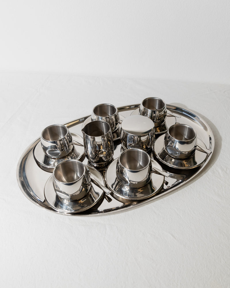 A polished metal tray showcases the Vintage Coffee Set - Italy 70s by Bottega Jacobs, featuring eight matching stainless steel cups and saucers arranged in two neat rows. The shiny surface reflects light, emphasizing the sleek, modern design of the set against a soft, white background.