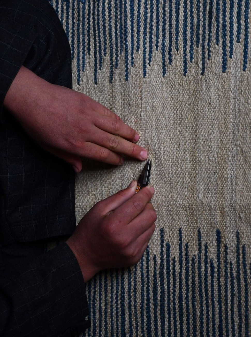 Two hands are seen repairing or trimming a handwoven wool kilim with a small tool. The fabric, adorned in natural dyes, features vertical lines in shades of blue and white. The hands are positioned close to the surface, indicating detailed, intricate work on this Navy Anar Kilim Runner - Handwoven Rug from ISHKAR.