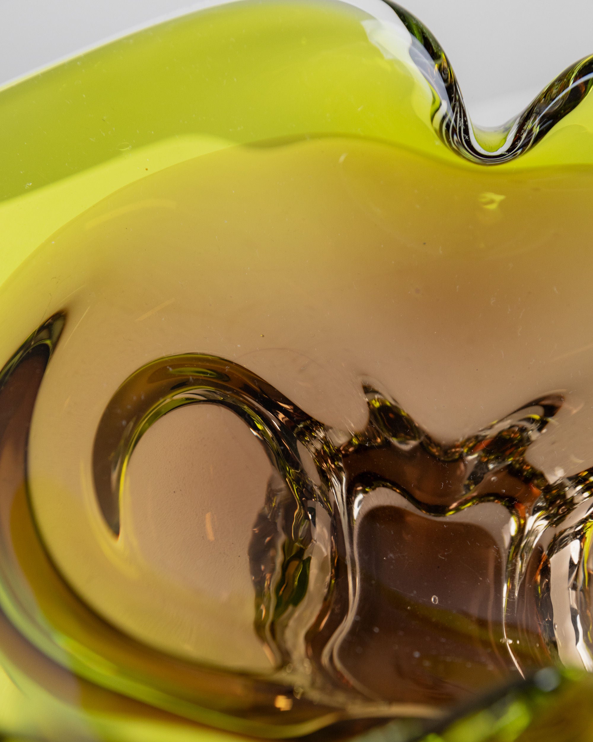 A close-up of the Bottega Jacobs Murano Glass Bowl from Italy, 1970s, reveals fluid, swirling shapes akin to vintage designs. It showcases a color gradient from green to amber, offering an abstract and dynamic look. The shiny surface reflects light gracefully, epitomizing Italian craftsmanship.
