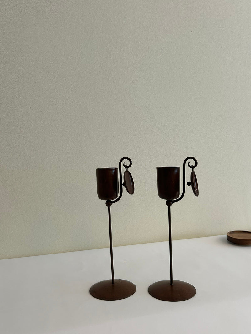 A pair of Vintage Brutalist Candle Holders by Flock Studio, featuring a curving arm and a small hanging ornament, stand on a white surface against a light green wall. These elegant metal holders with decorative elements enhance their sculptural aesthetic.