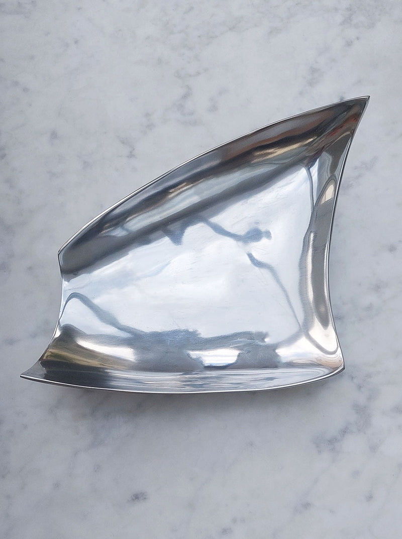A reflective, polished Les Objoies Brass 80's Large Tray shaped like an abstract, curved leaf placed on a grey marble surface, serving as elegant home decor.