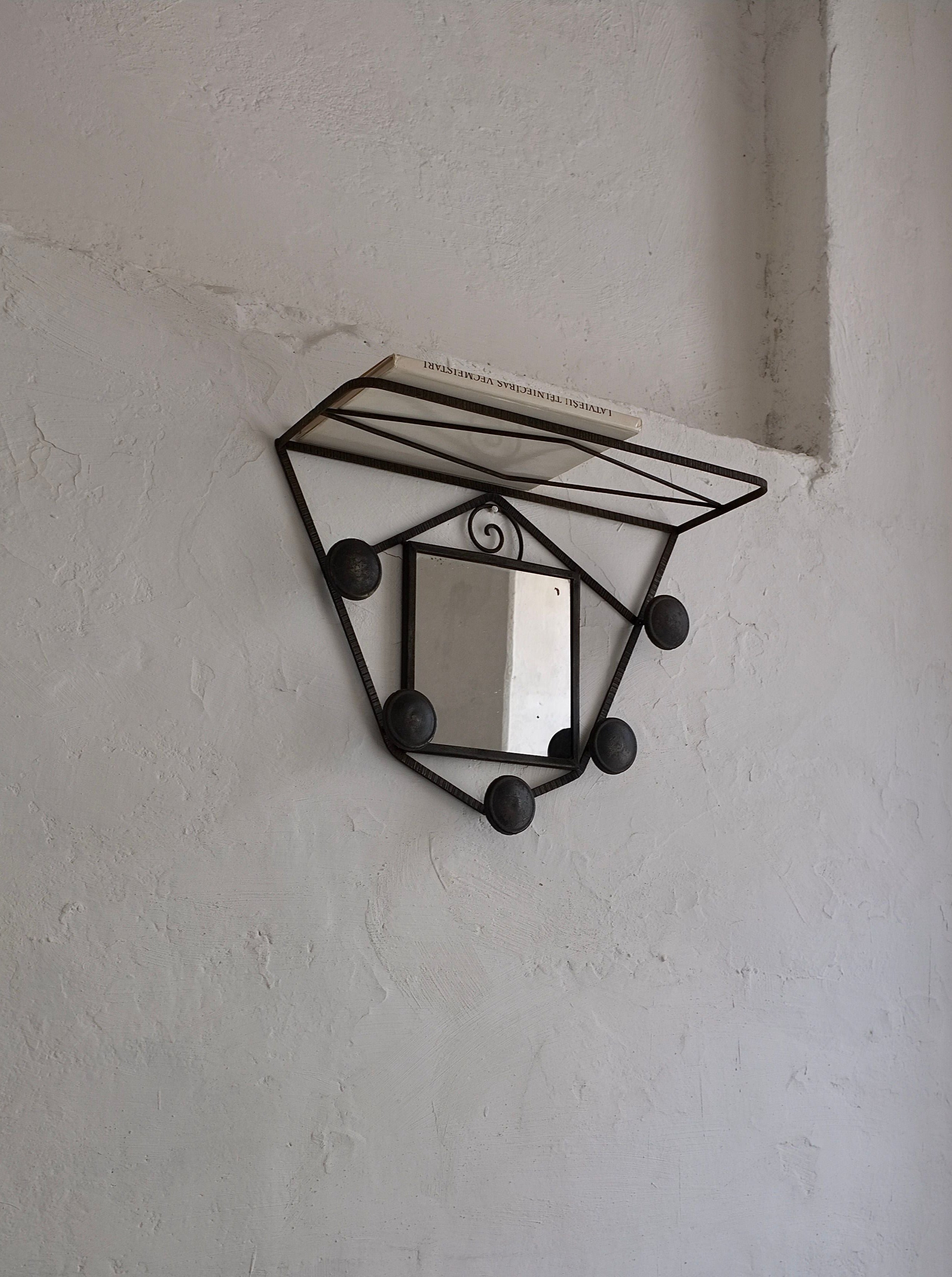 A geometric metal wall shelf with a diamond-shaped frame is mounted on a textured, white wall. This vintage entrance wall unit by Veter Vintage from the 1930s France collection—an Art Deco Wrought Iron Wall Mirror Coat Rack—holds a book with a white cover. Below it, the rectangular mirror attached reflects a portion of the room.