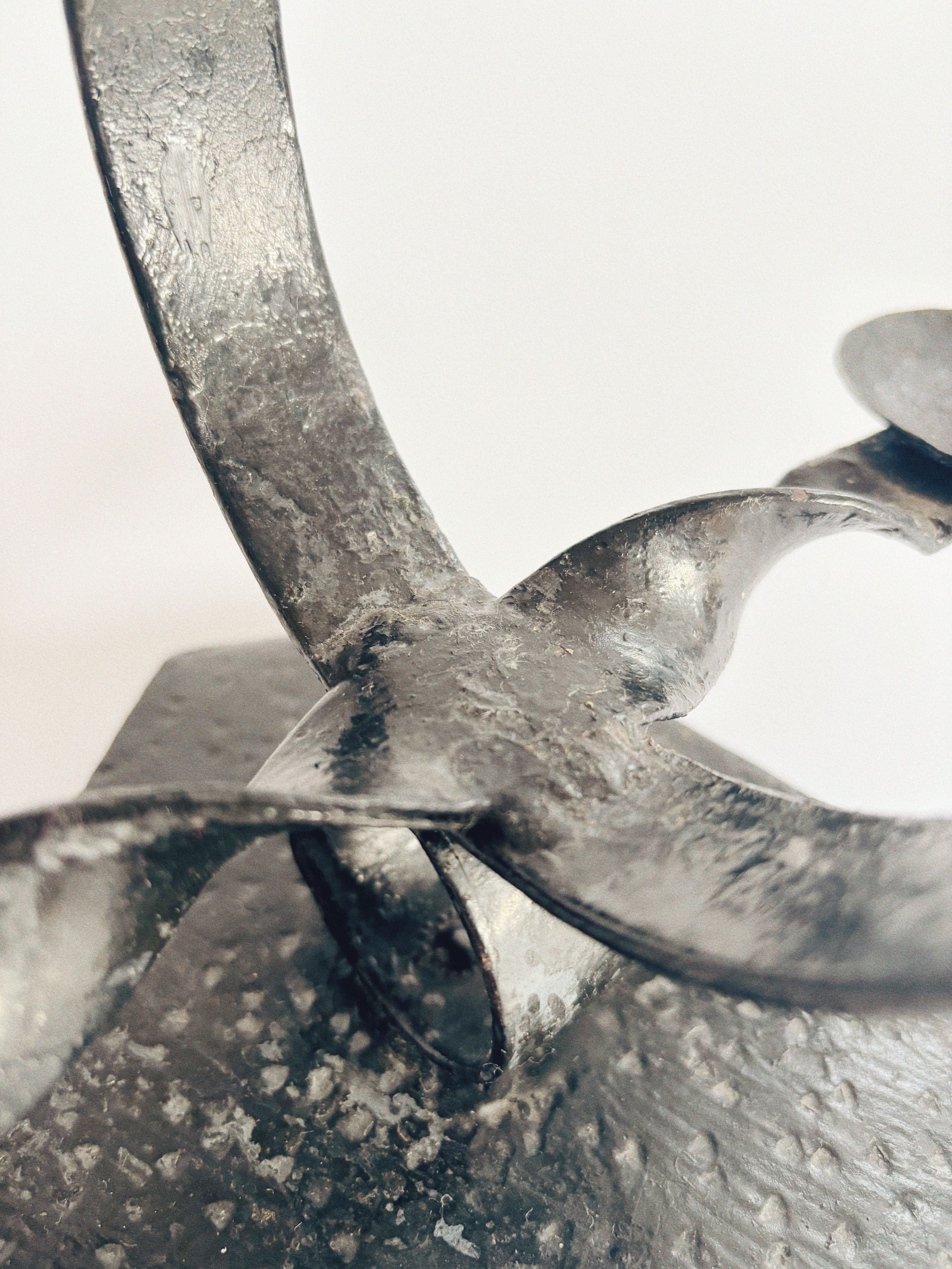 A close-up of the Black Metal Candlestick by Huerto reveals its twisted, ribbon-style arms forming overlapping loops. The metallic surface is textured and weathered, reminiscent of vintage artistry, set against a plain background.