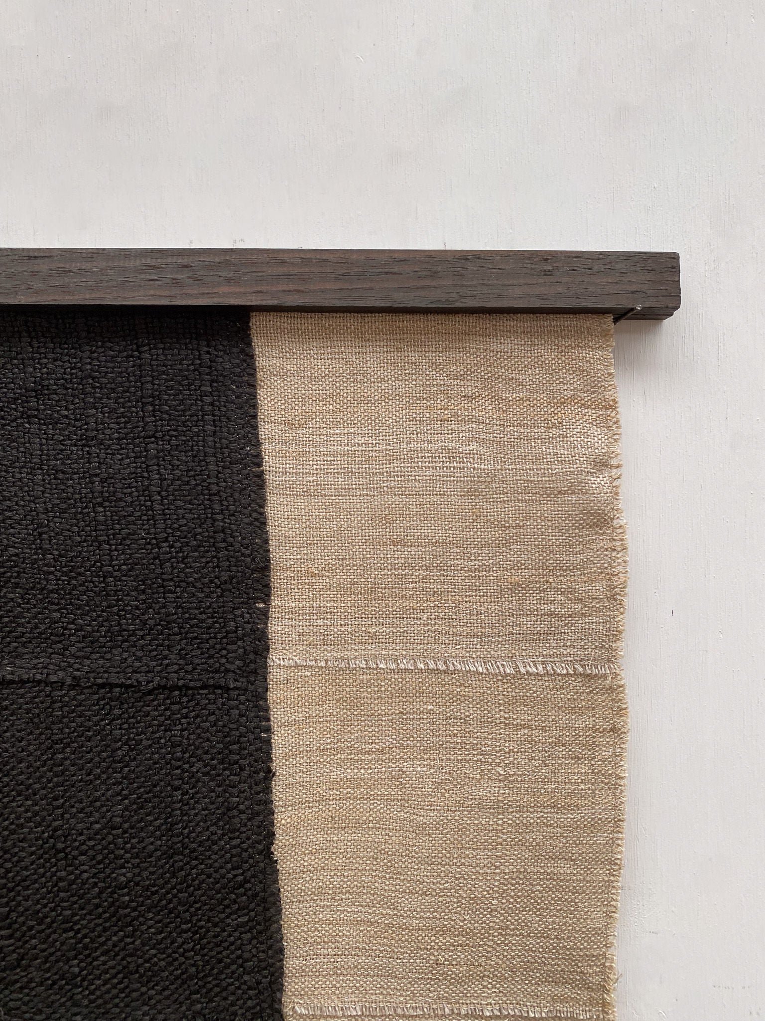 The artwork "Paths" by Sundance Studio is a woven textile crafted from antique cotton. It features two vertical sections, one black and the other beige, elegantly mounted on a dark wooden rod against a light wall.