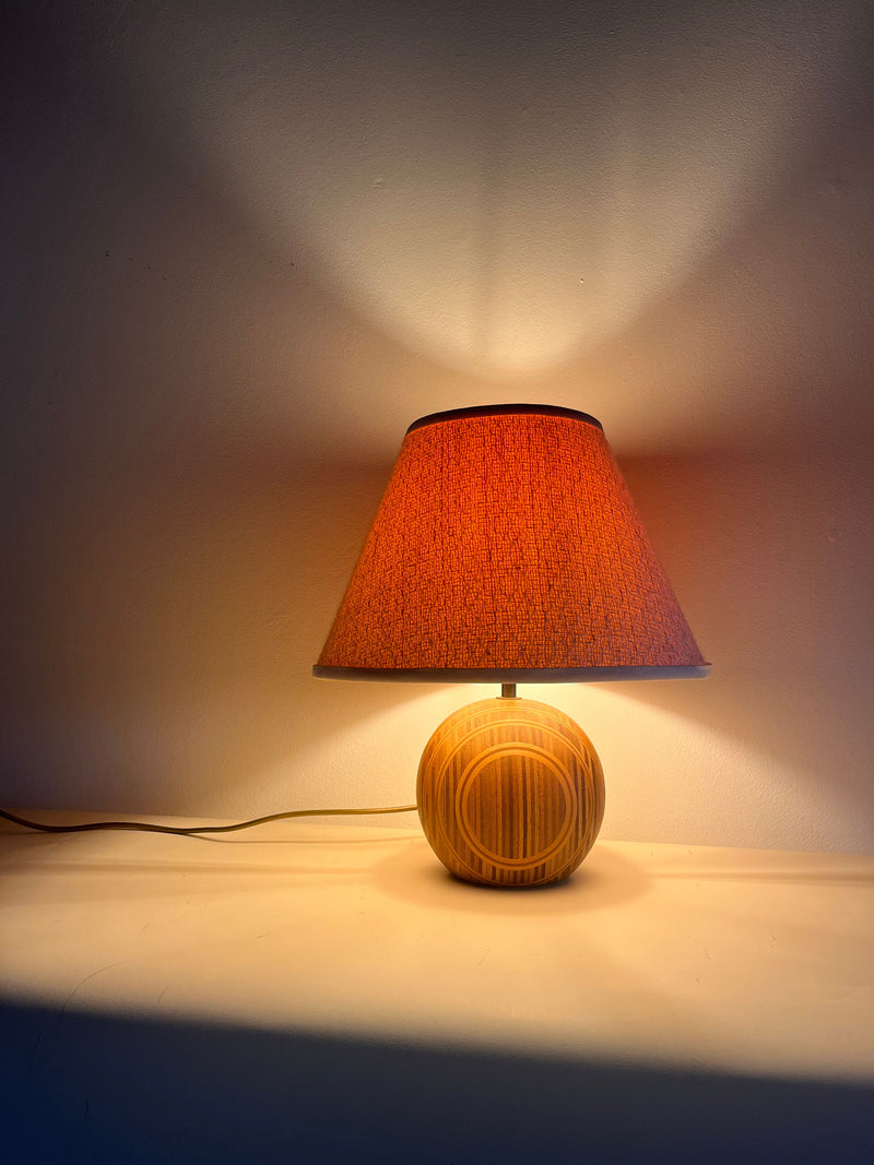 A Wood Table Lamp 1970s by Maud Vaughan featuring a textured wooden base and a red-orange boucle shade is placed on a white surface, casting a soft glow and creating intricate light and shadow patterns on the neutral-colored wall behind it.