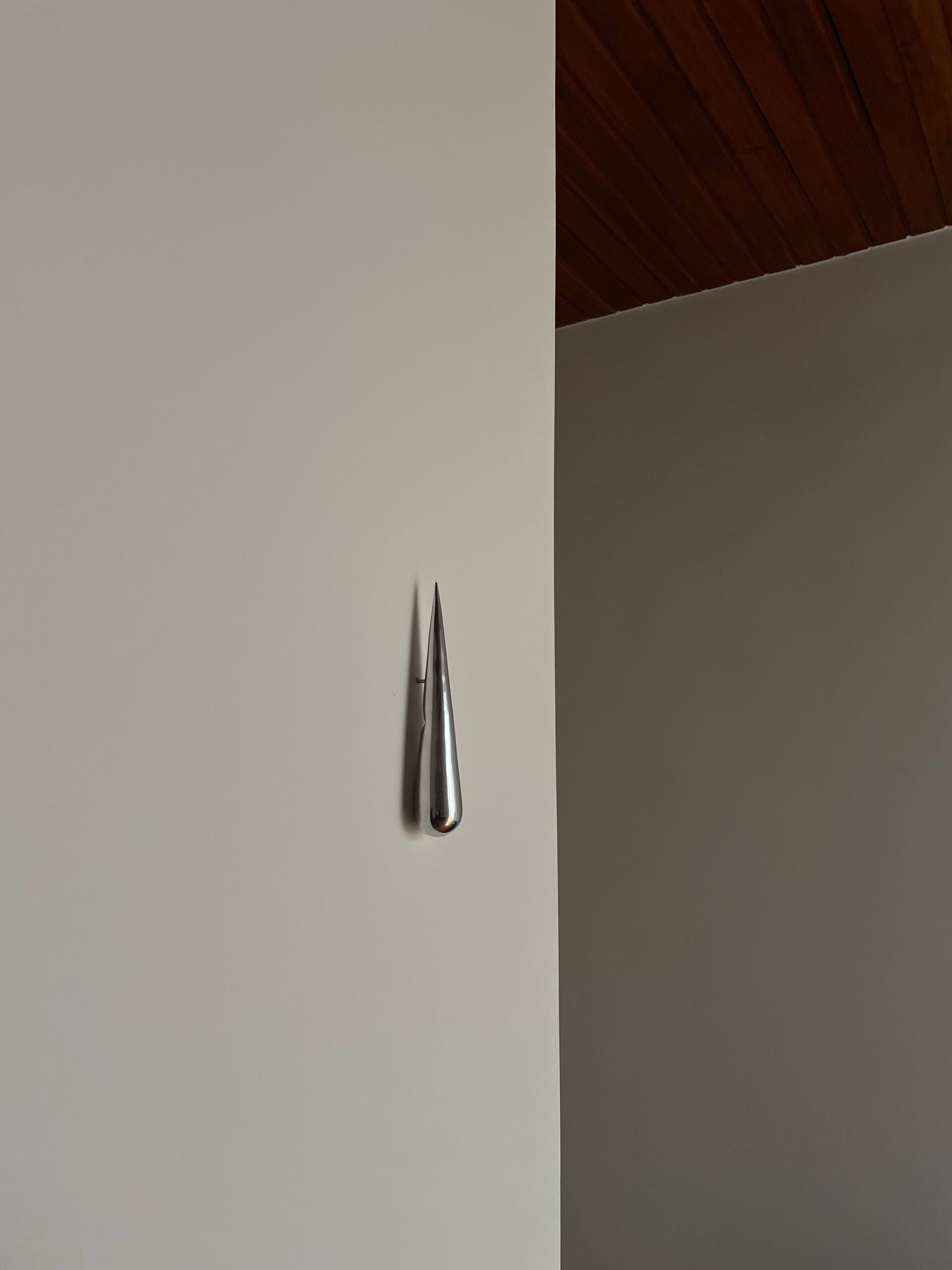 The "GOTA" Bottle Opener by Pedro Avila is a sleek, metallic, droplet-shaped object crafted from cast aluminum. It is mounted vertically on a minimalist, light-colored wall, with a background of wooden-paneled ceilings that enhances the clean, modern aesthetic of this limited-edition piece.