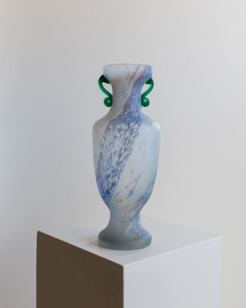 The Murano Glass Vase 70s by Bottega Jacobs features a translucent body with a swirling blue and white pattern, accented by two green, curved handles. It's displayed on a pedestal with a softly lit, neutral-toned background, evoking the elegance of an XL vintage Murano piece.