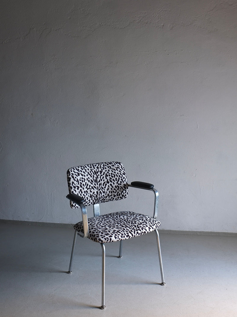 A single Veter Vintage Minimalist Animal Print Chrome Desk Arm Chair | Sweden | 1960s is positioned against a plain, textured, light-colored wall. The chair has metal legs and leather armrests, standing on a smooth, light grey floor. The setting appears minimalist and empty.
