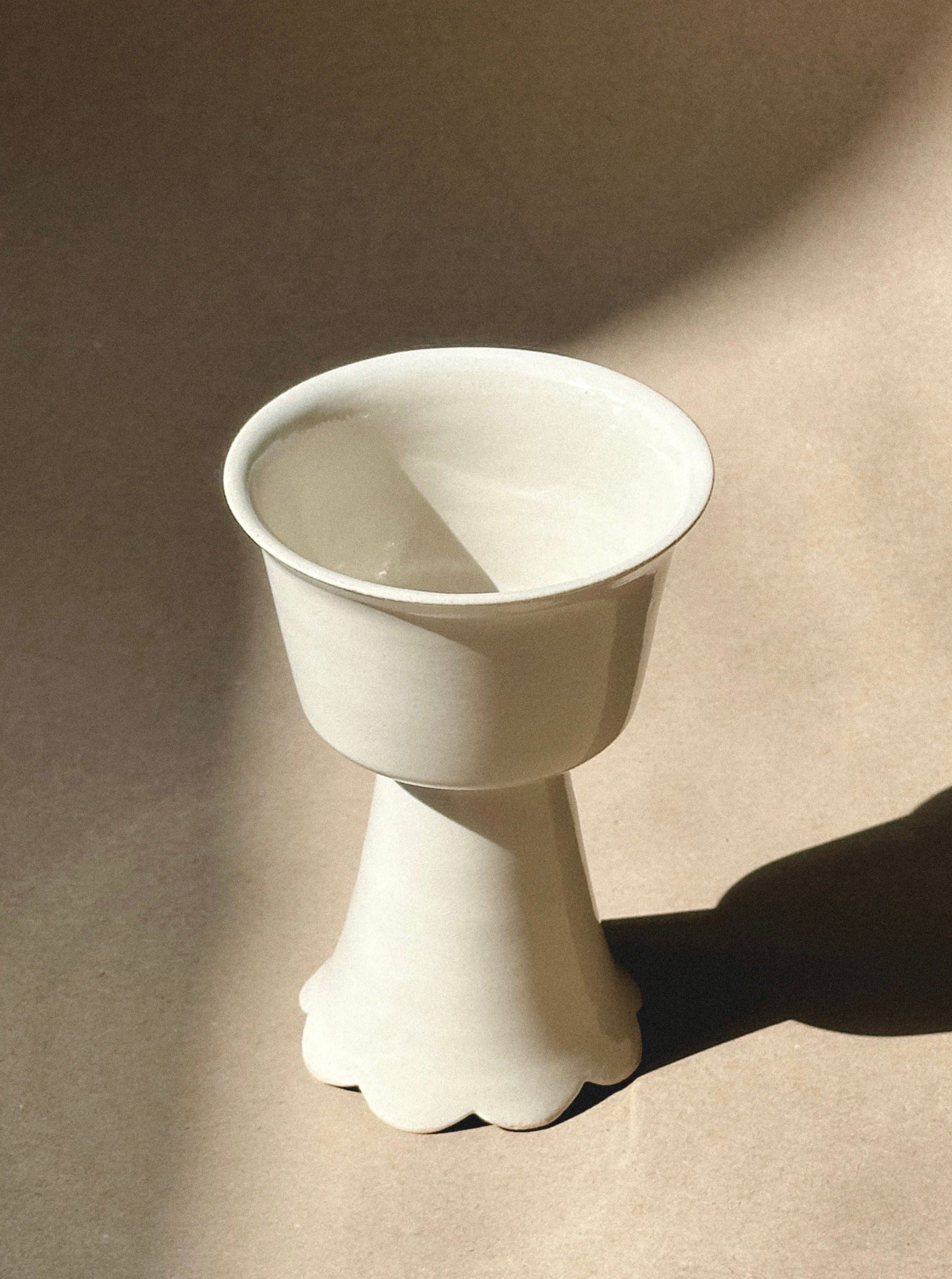 A Flowery Wine Goblet with a unique design, featuring a wide, shallow bowl on top of a flared, pedestal-like base, reminiscent of elegant wine goblets. The vase is placed on a beige surface with soft shadows cast by sunlight. The overall color of this handmade stoneware piece by noki ceramics is a creamy white.