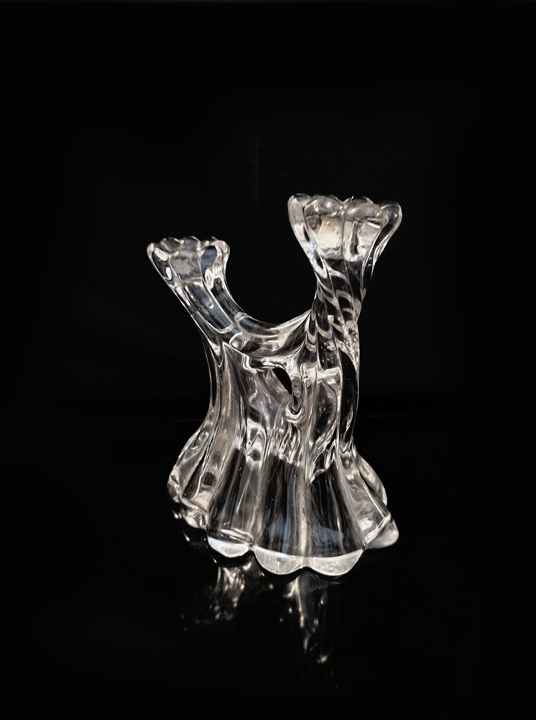 The Glass Candle Holder by Les Objoies, featuring a modern and artistic design with fluid, twisting shapes, stands out against a black background for a striking contrast. Its organic form captures and reflects light beautifully, highlighting its intricate design and delicate craftsmanship.