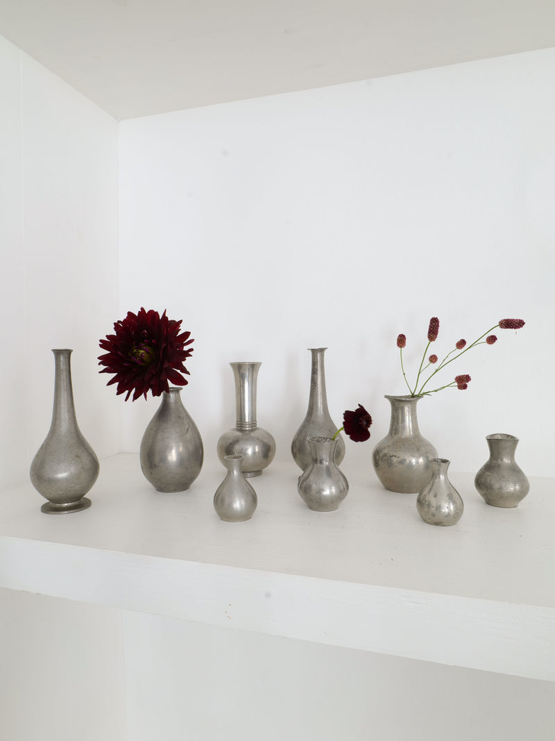 Displayed on a white shelf is the Just Andersen Mini Vase Collection by Collection apart, featuring various silver vases in different shapes and sizes. Some of these miniature vases hold dark red flowers and small foliage stems, creating a striking contrast against the minimalist white background.