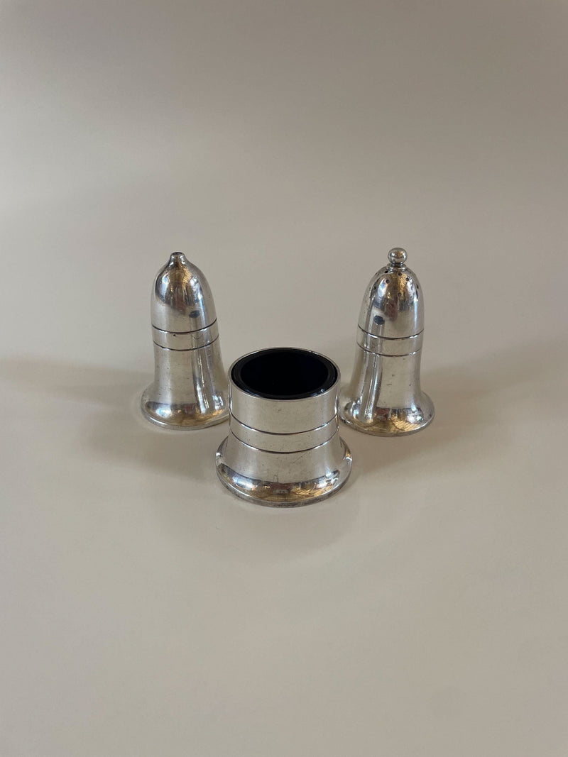 Three small silver-colored metal objects are arranged on a flat surface. Two objects resemble traditional bell-shaped pepper shakers, while the third is shorter and cylindrical with an open top, possibly a napkin ring or holder. This setup evokes the charm of a Vintage Mappin and Webb Cruet Set by Maud Vaughan.