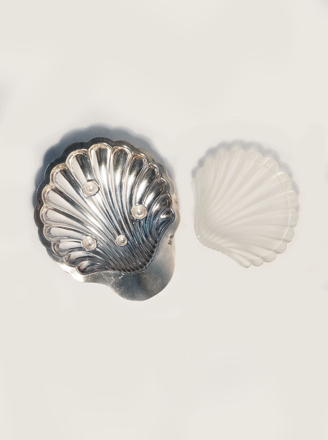 A silver-plated, scallop-shaped Shell Butter Dish by Les Objoies, featuring a ribbed pattern and adorned with pearls and frosted glass, sits elegantly on a light background. The larger metallic dish reflects light beautifully, while the glass dish appears delicately translucent.