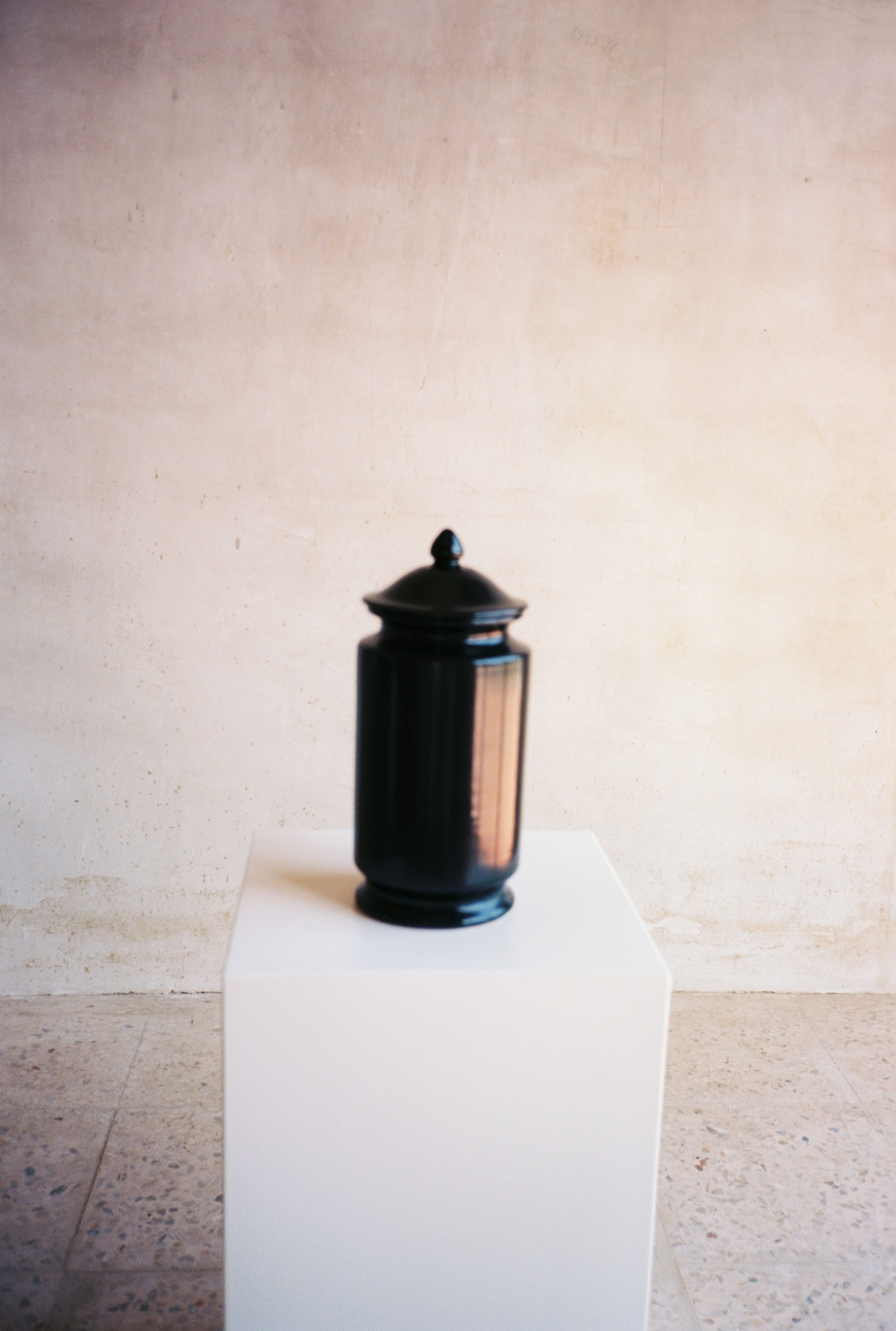 The COSEINCORSO "Albarello" Jar in matte black sits on a white pedestal against a textured light wall, creating a minimalist display.