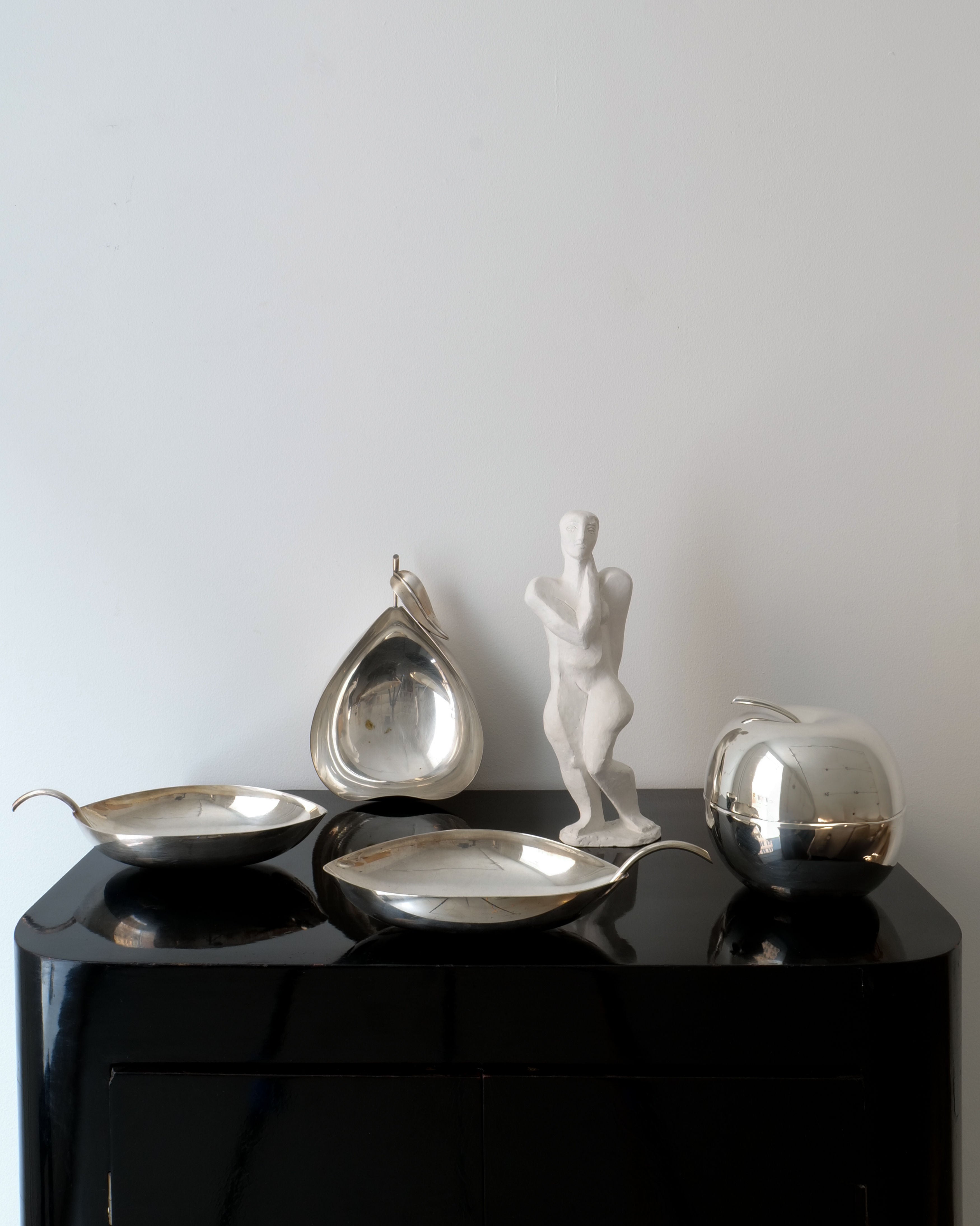 A Collection apart black cabinet displays silver trays, a pear-shaped dish, and the Christofle Apple Jar Lino Sabattini with its polished modernist design. Next to these is an abstract white sculpture of a human figure by Christofle, reminiscent of Lino Sabattini's sleek creations. The scene is set against a plain white wall.