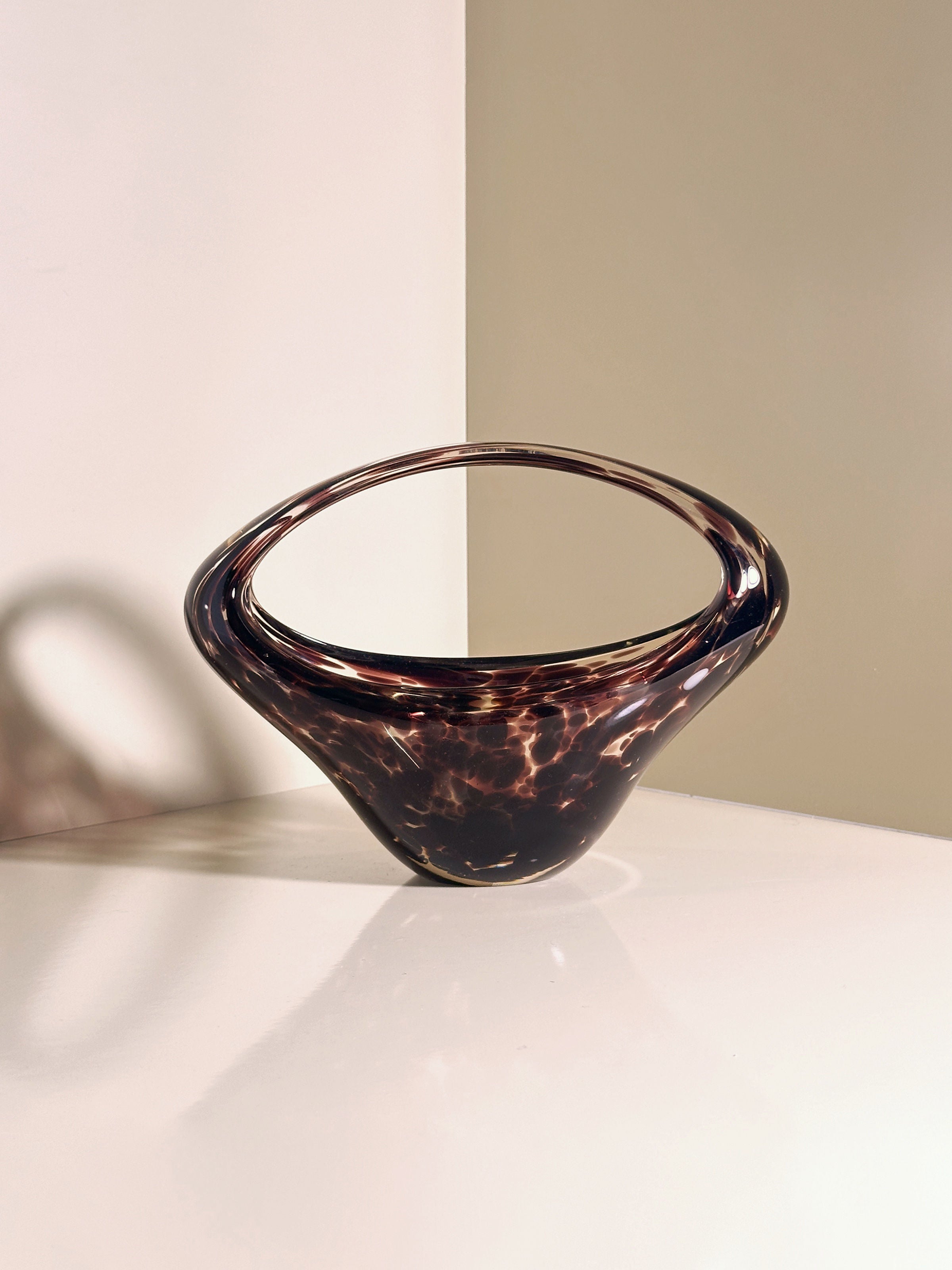 The Murano Tortoise Glass Handle Bowl - 1970s by Dodo Vintage, with its sleek design and elliptical opening, features a marbled pattern and reflects softly on a white surface, casting shadows against a neutral wall reminiscent of chic 1970s style.
