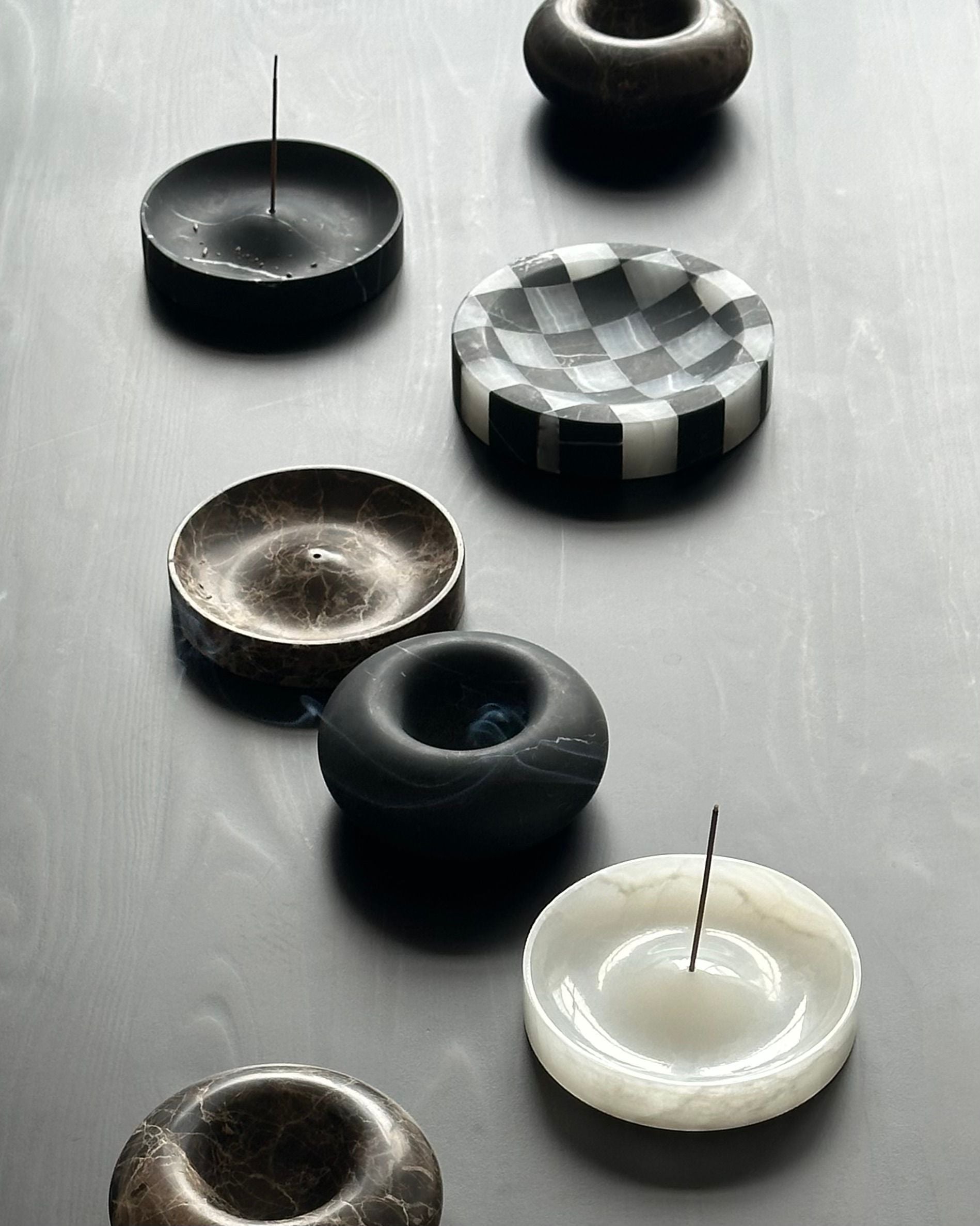 CARRARA Studio's 'ALTAR' Incense Holders—handcrafted, round marble pieces in diverse colors and patterns—are shown on a smooth, dark surface, some holding incense sticks.