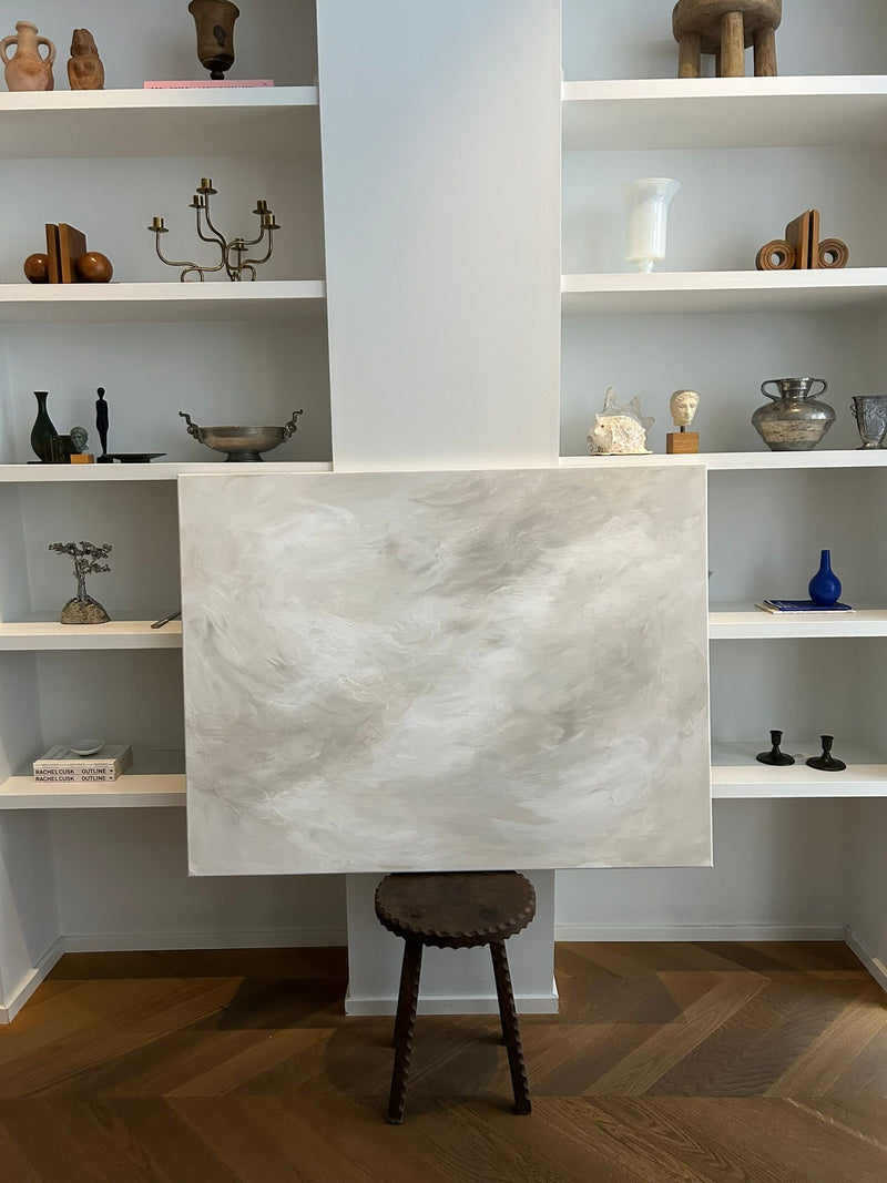 The "Celestial Drift" painting by Mattias Storm, featuring soft gray and white swirls, sits on a wooden stool in front of shelves showcasing decorative items like vases, sculptures, and books. The flooring is wooden.