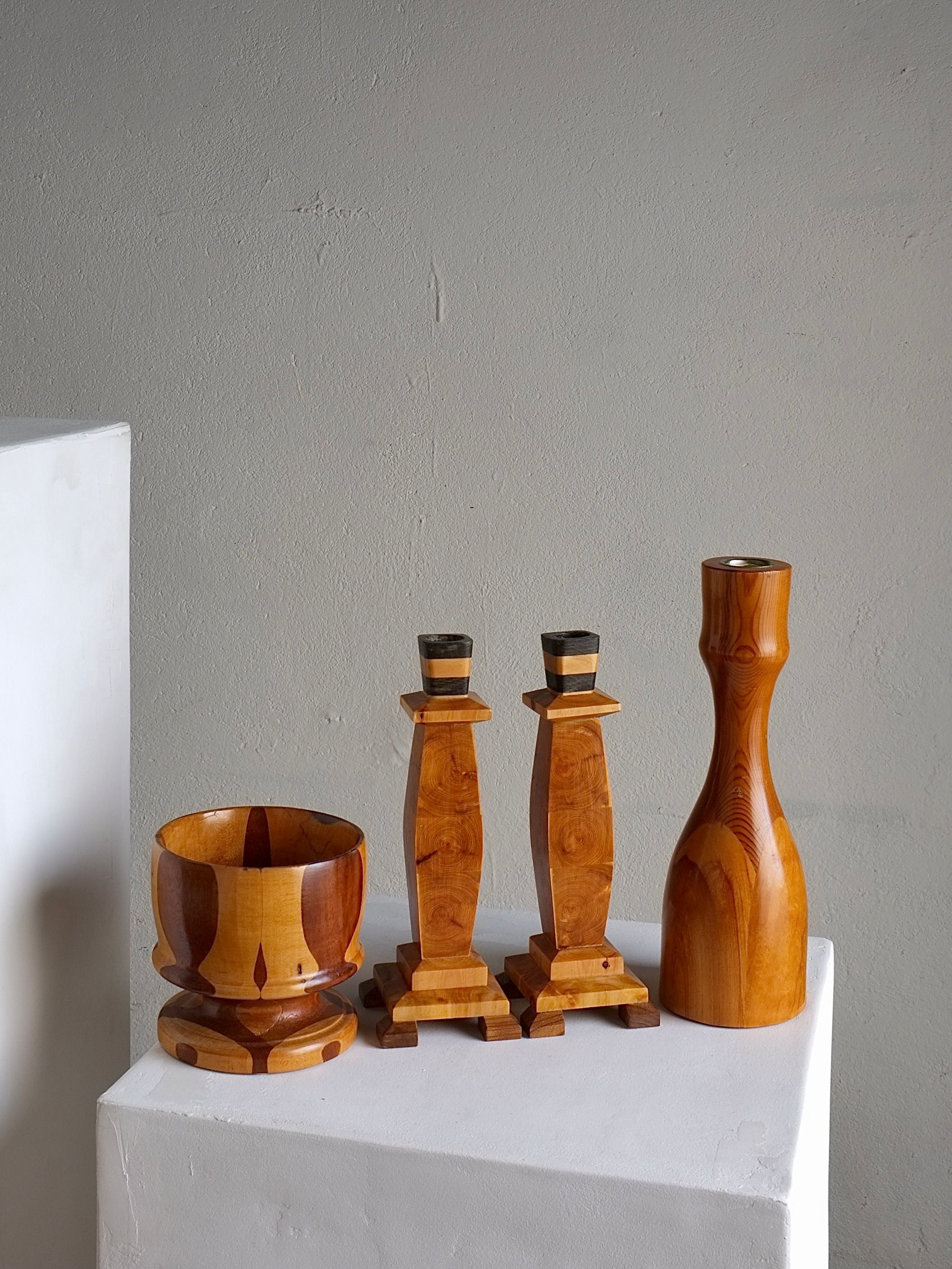 A photograph featuring the Veter Vintage Carved Wooden Candle Holders and Bowl Set from Sweden, including a wooden bowl, two candlestick holders, and a vase arranged on two white pedestals against a gray background. The items exhibit light and dark wood tones from the 1970s, showcasing intricate craftsmanship.