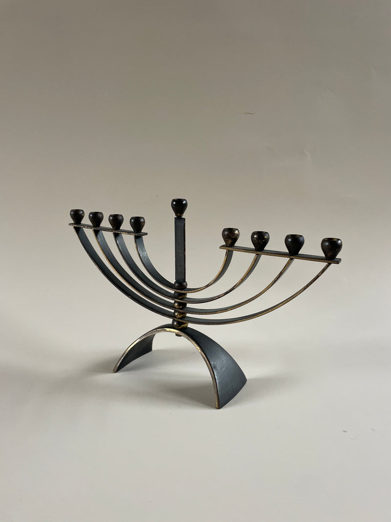 The Maud Vaughan Menorah Candelabra is a contemporary black and gold metal menorah featuring eight branches with a central elevated branch on a curved base, perfect for slim taper candles, set against a neutral background.