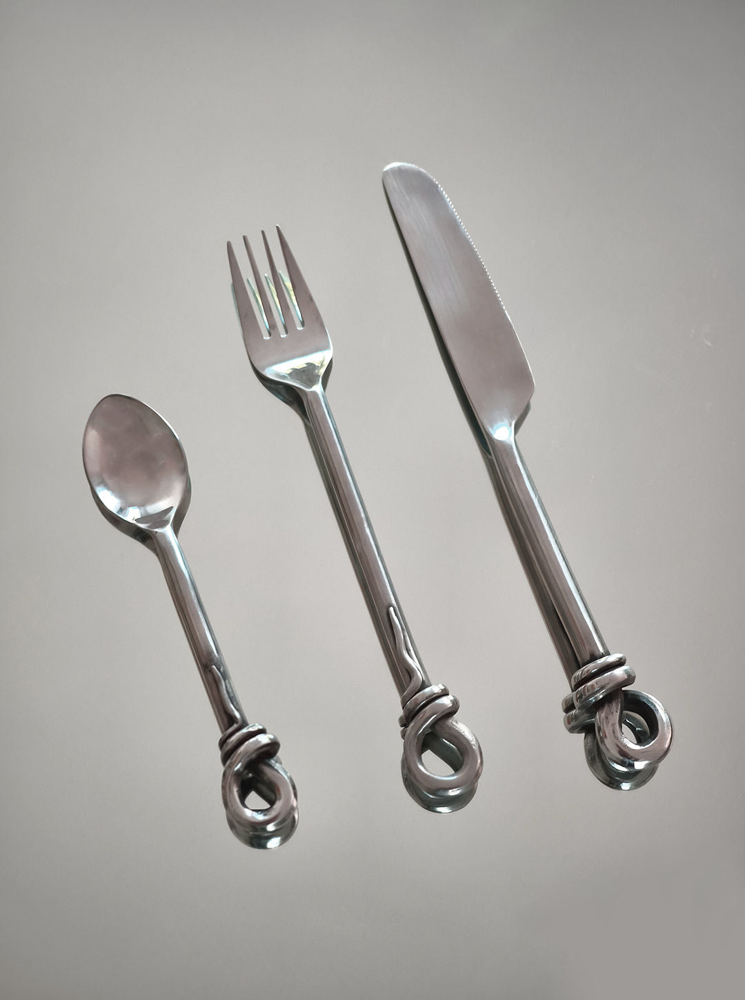 A Set of 3 twist cutlery from Les Objoies, featuring a shiny silver spoon, fork, and knife with distinctive twist vintage handles, arranged in a row on a gray background.