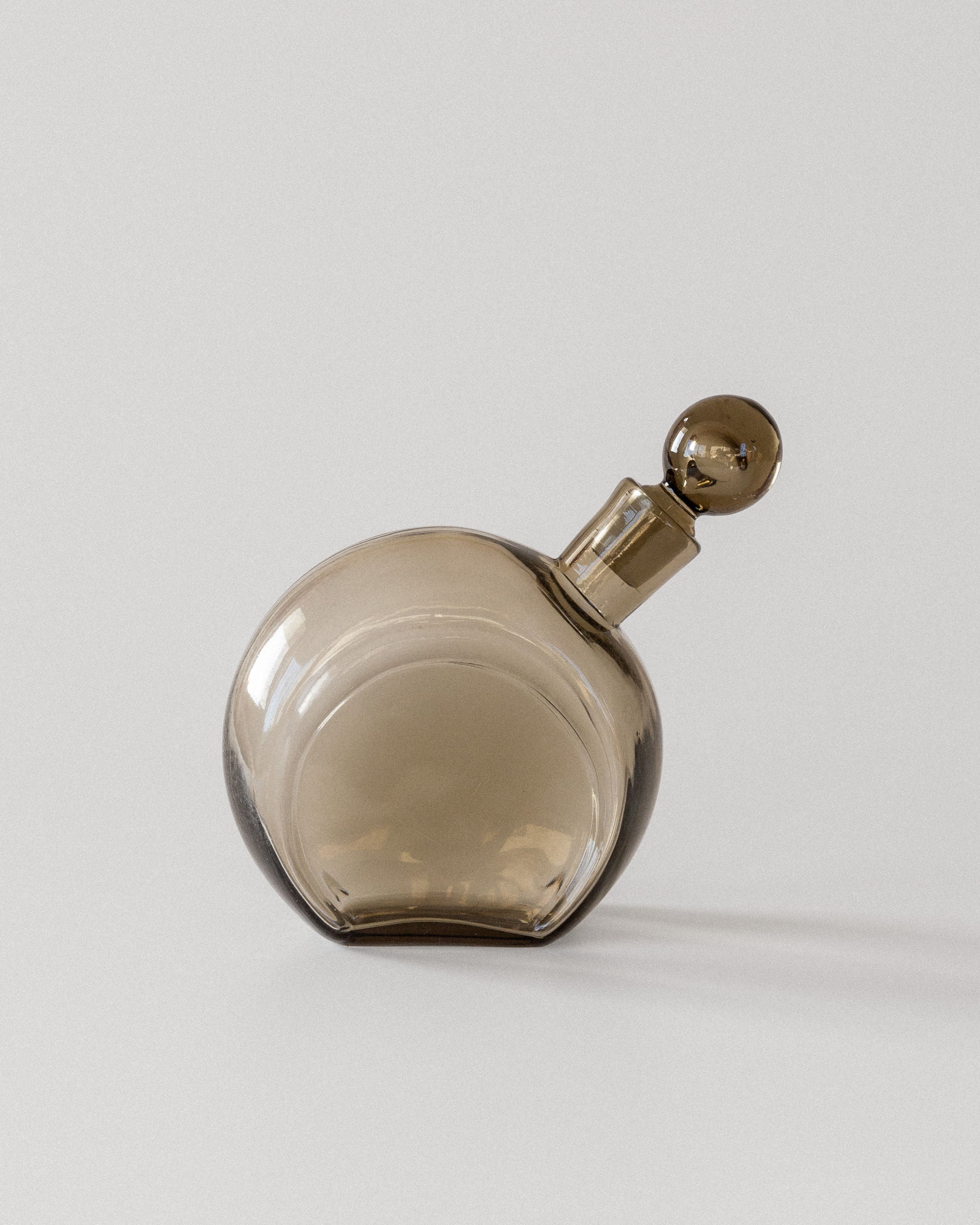 A 1930s Glass Decanter by Harald Notini for Spigel, featuring smoky brown glass and a spherical stopper, rests elegantly on its side against a white background. The tinted glass casts a gentle shadow, reflecting Swedish modernist design.