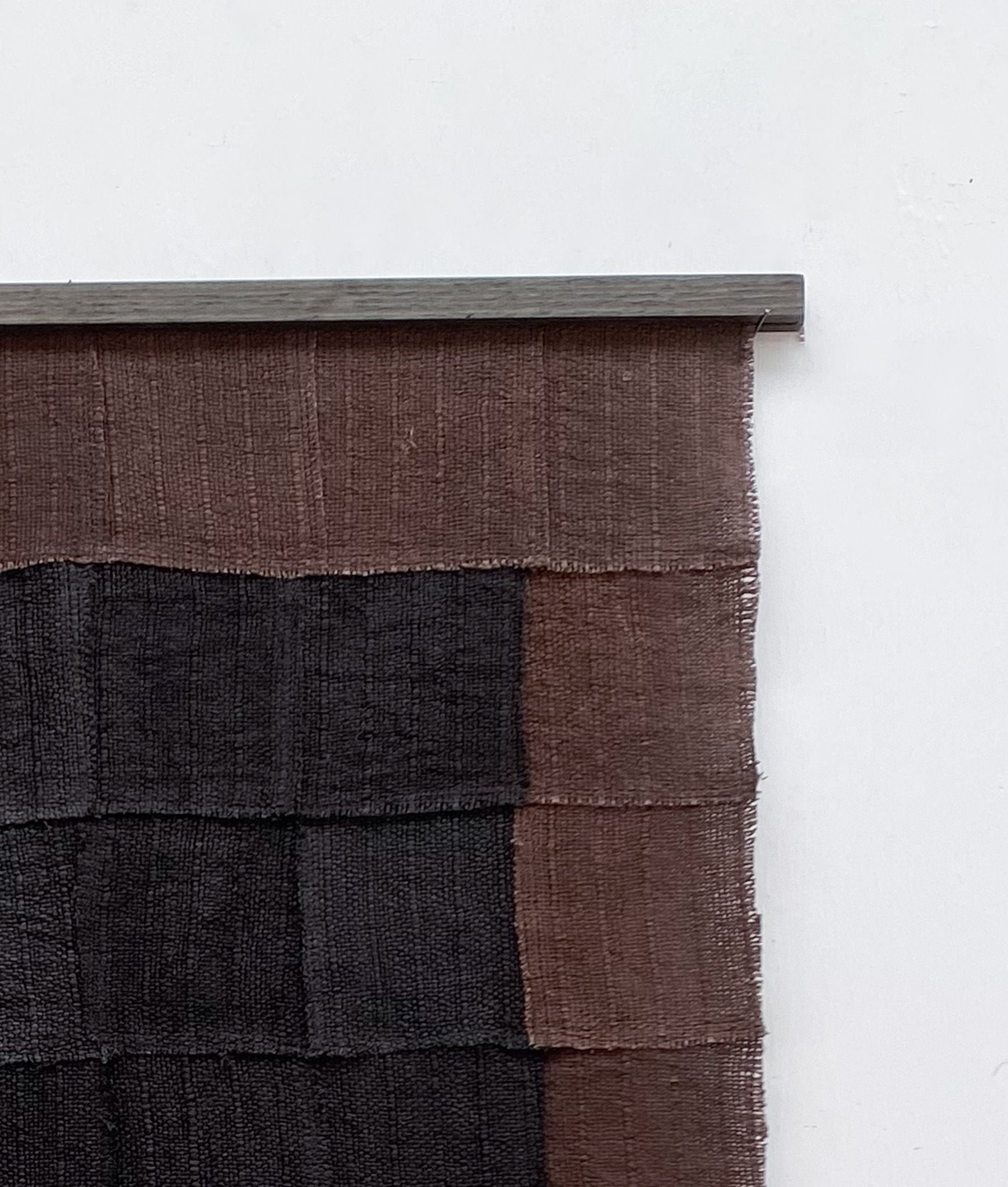 The Sundance Studio's Ground Echo, a textile wall hanging from the Boketto residency collection, features a grid pattern of dark brown and black squares reflecting the sweeping plains of Yaegl Country. It hangs gracefully from a simple rod against a plain white backdrop.
