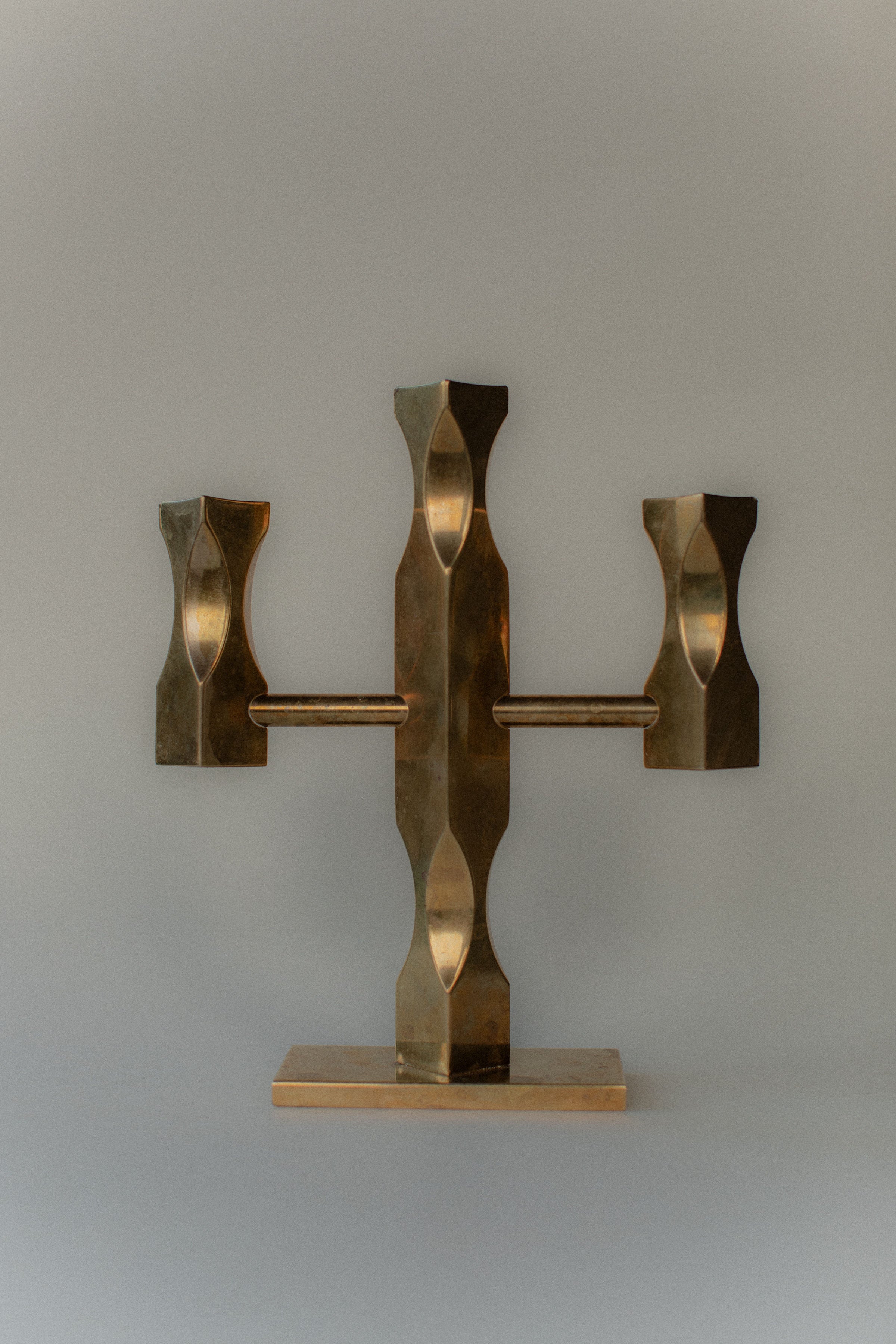 The Brass Candleholder by Out For Lunch is a bronze-colored metal sculpture with a Brutalist style, featuring abstract geometric shapes. It has a central vertical piece, two symmetrical horizontal extensions on a flat base, and is set against a plain background.