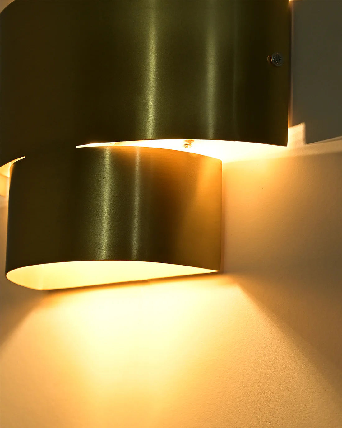 The Aura Wall Sconce by LMNOH is a modern brass fixture with a metallic finish, emitting warm ambient light on a neutral wall. It casts soft shadows that accentuate its sleek design, making it ideal for contemporary interiors.